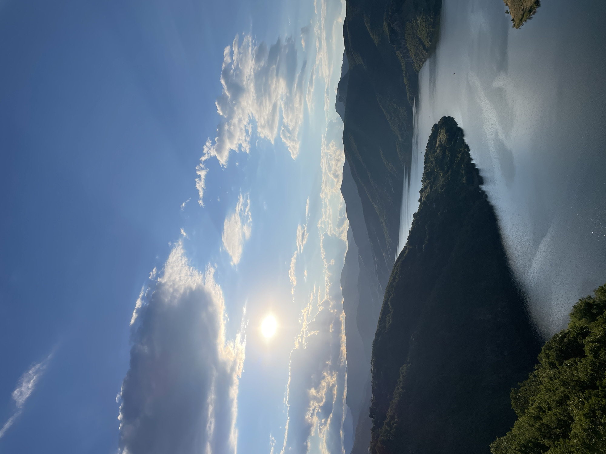 This is a landscape photograph taken from an elevated viewpoint overlooking a large body of water, likely a reservoir, surrounded by rolling, tree-covered mountains. The sun is shining brightly, partially obscured by dramatic, scattered clouds, casting rays of light that illuminate the scene. The water's surface reflects the sky and surrounding landscape. The mountains recede into the distance, with layered peaks creating a sense of depth. The foreground features a dense, green forest on the left side of the frame. The overall impression is one of natural beauty and tranquility, likely captured during late afternoon or early evening due to the quality of light. The scene is consistent with the topography and appearance of the area around the dam and the dam itself in Borovitsa, Bulgaria.