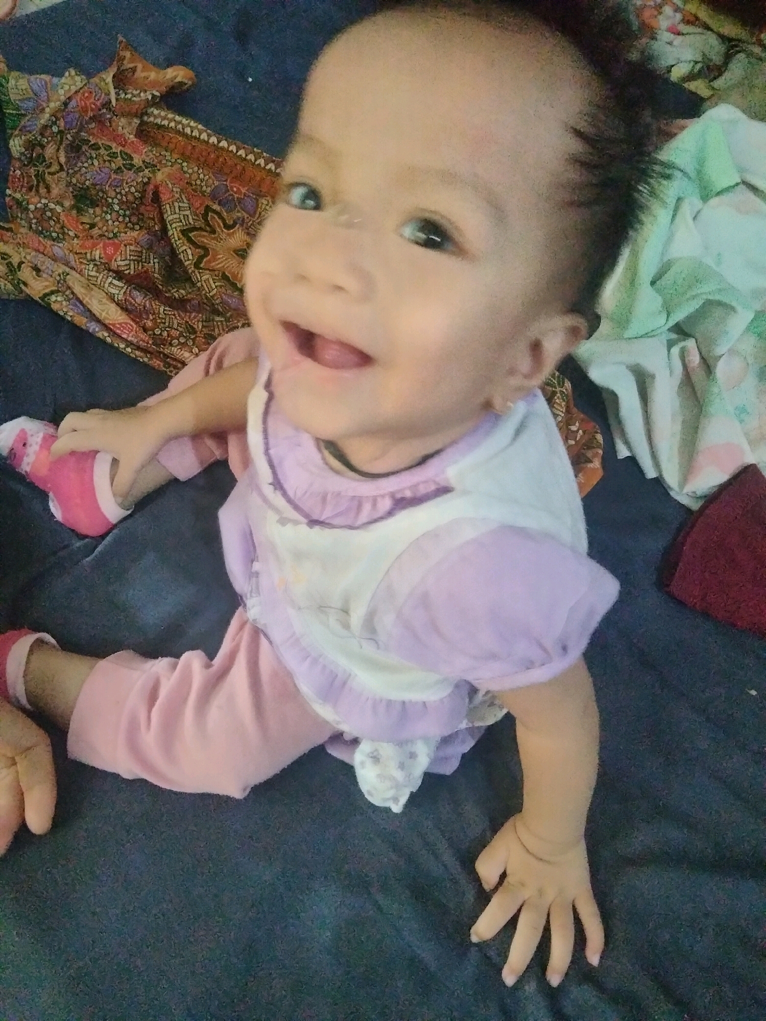 A baby is sitting on a dark blue blanket, likely indoors in Suka Sari, Indonesia. The baby is facing the camera with a wide smile, showing its tongue. They are wearing a white and lavender shirt and pink pants and socks. To the left of the baby is a colorful fabric with a batik pattern. To the right are other pieces of fabric, one light green and white with a floral pattern, and a dark red cloth. The baby is positioned in the center of the frame, with its legs bent and arms reaching out. The lighting suggests it may be daytime.