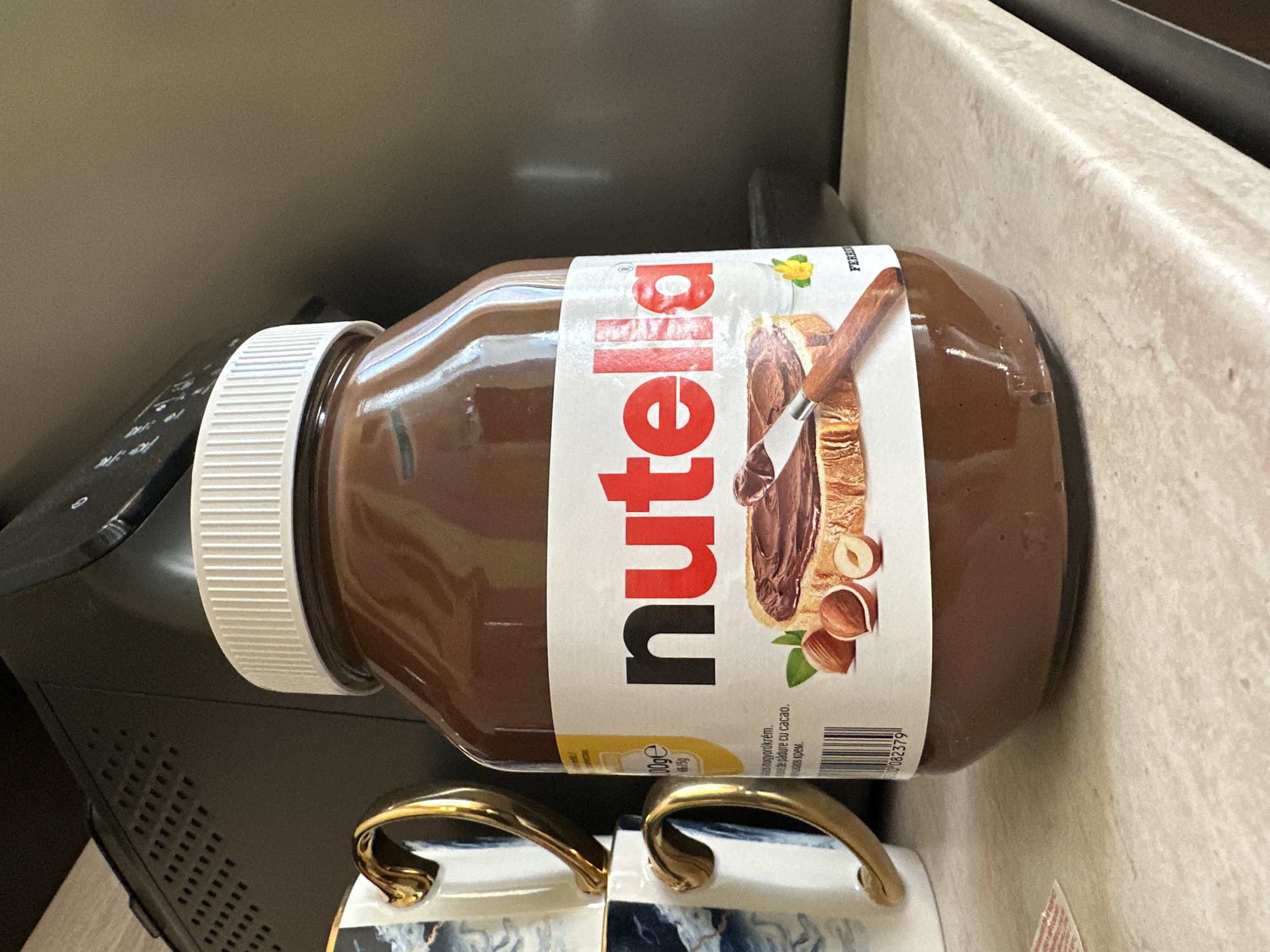 A close-up, eye-level shot shows a large jar of Nutella positioned on a light-colored countertop. The jar is the dominant subject, with its iconic label clearly visible. The label features the "Nutella" logo in bold red and black lettering, along with an illustration of a slice of bread spread with Nutella, hazelnuts, and a knife. Text in various languages, including Hungarian and what appears to be Romanian, is visible on the label, along with a barcode.
To the left of the Nutella jar, two white ceramic mugs with gold handles are stacked. The mugs have a blue and white marbled pattern. In the background, a dark, textured object, likely an electronic device, is visible. The countertop itself has a speckled, marble-like pattern. The lighting is bright, suggesting an indoor environment with artificial or natural light sources. The scene is static, with no visible people or animals.
