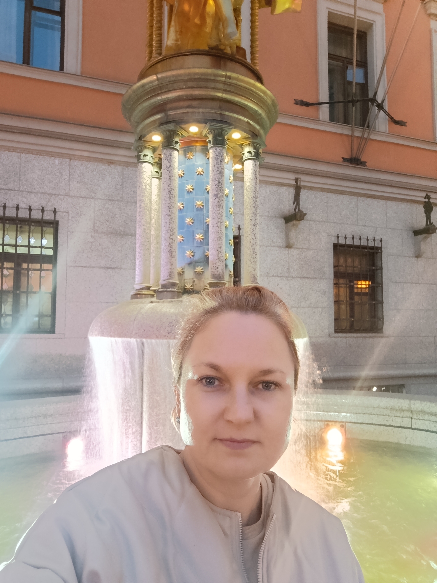 A woman with blonde hair tied back is taking a selfie in front of a fountain in Moscow, Russia. She is wearing a light-colored jacket. The fountain has decorative columns with star-shaped accents and is illuminated from within. Behind the fountain, a building with orange-colored walls and windows can be seen. The fountain's water is flowing and reflecting the colorful lights. The scene appears to be outdoors at night or in low light conditions.