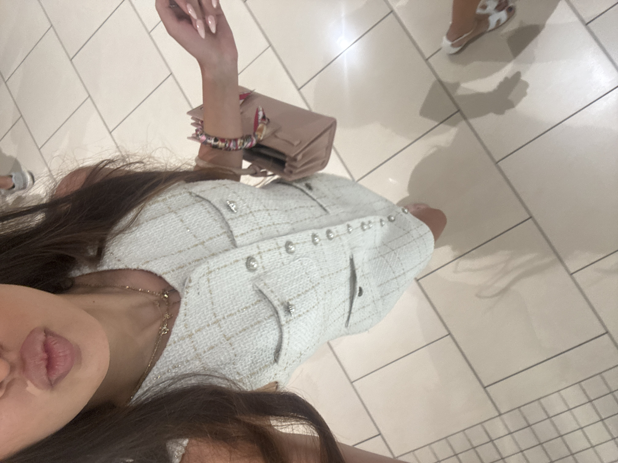A woman, viewed from a high angle and slightly from above, stands in an indoor setting with beige tiled flooring. She is wearing a sleeveless, light-colored tweed dress with silver buttons and two pockets on the chest. She has long, dark hair and is wearing a necklace. Her right hand, adorned with manicured nails, is visible, holding a small, light pink purse. A colorful bracelet is on her left wrist. Her lips are pursed. Her feet are not fully visible, but one foot is wearing a white sandal with a patterned strap. In the background, another person's legs and feet are visible, wearing grey sneakers. The environment appears to be a well-lit, public space, possibly a shopping mall or a similar retail environment in Aventura, Florida.