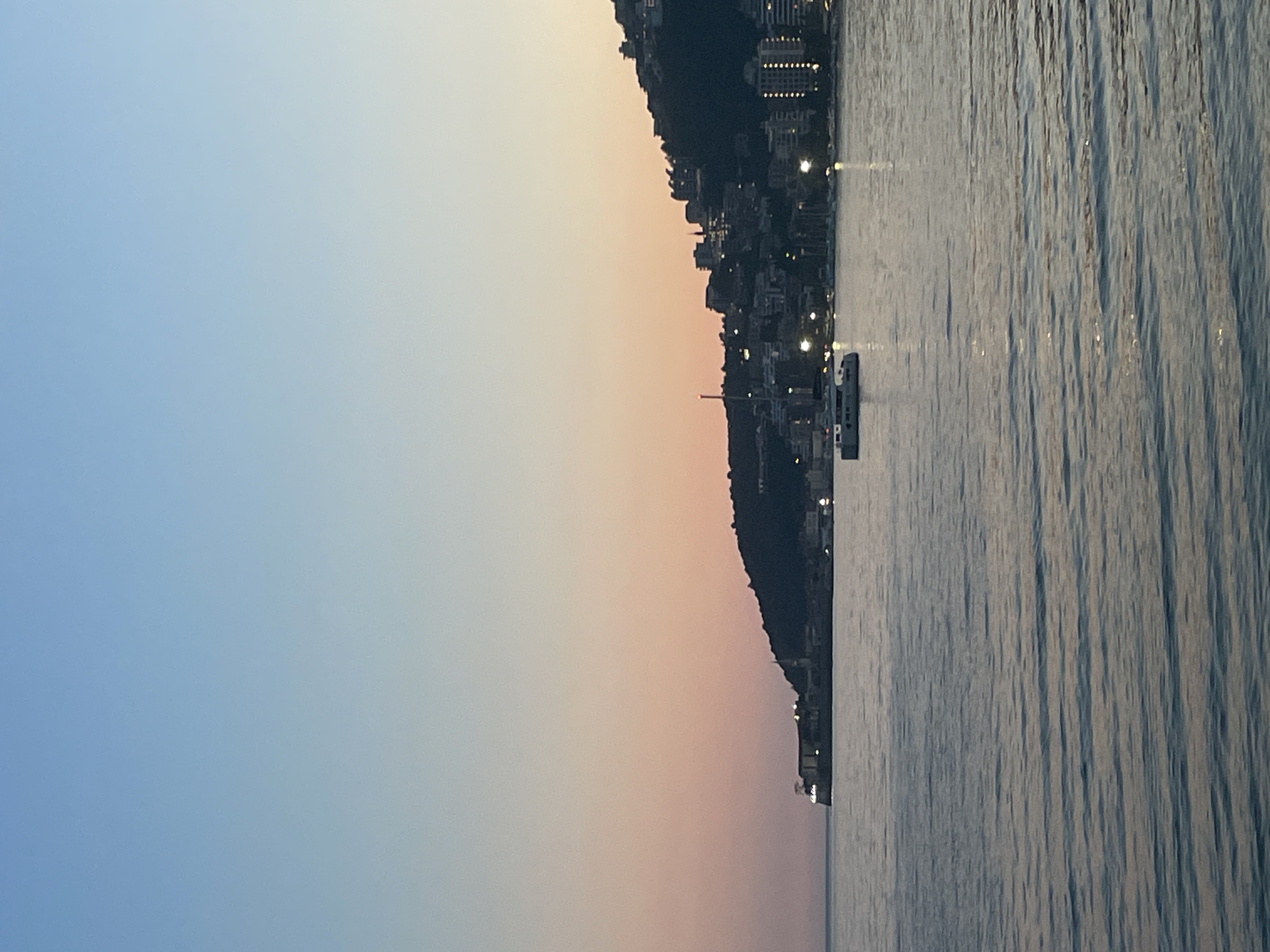 The image shows the coastline of Budva, Montenegro at dusk or dawn. The sky transitions from blue to peach/pink. A hillside with buildings and lights runs along the middle of the image, where it borders the water. The water has a textured surface, reflecting light. A small white boat is visible in the water near the hillside.