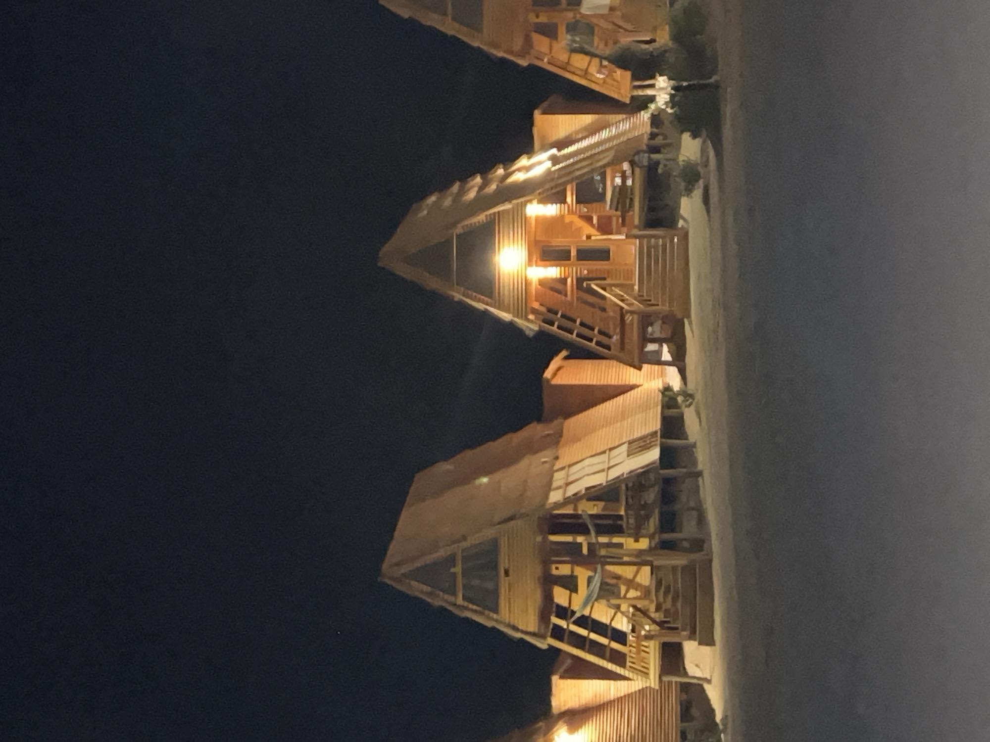 The image shows a nighttime view of several A-frame wooden cabins located on a sandy area. The cabins are illuminated from within, casting a warm glow. The sky is dark and clear, with a few faint stars visible. The foreground consists of a sandy ground with some sparse vegetation. The cabins appear to be part of a resort or accommodation facility in Nueiba, Egypt, likely situated near the coast. There are no people visible in the image, and the scene is still and quiet. The architecture of the cabins is distinctive, with steep pitched roofs and wooden exteriors, typical of some tourist accommodations in coastal or natural environments.