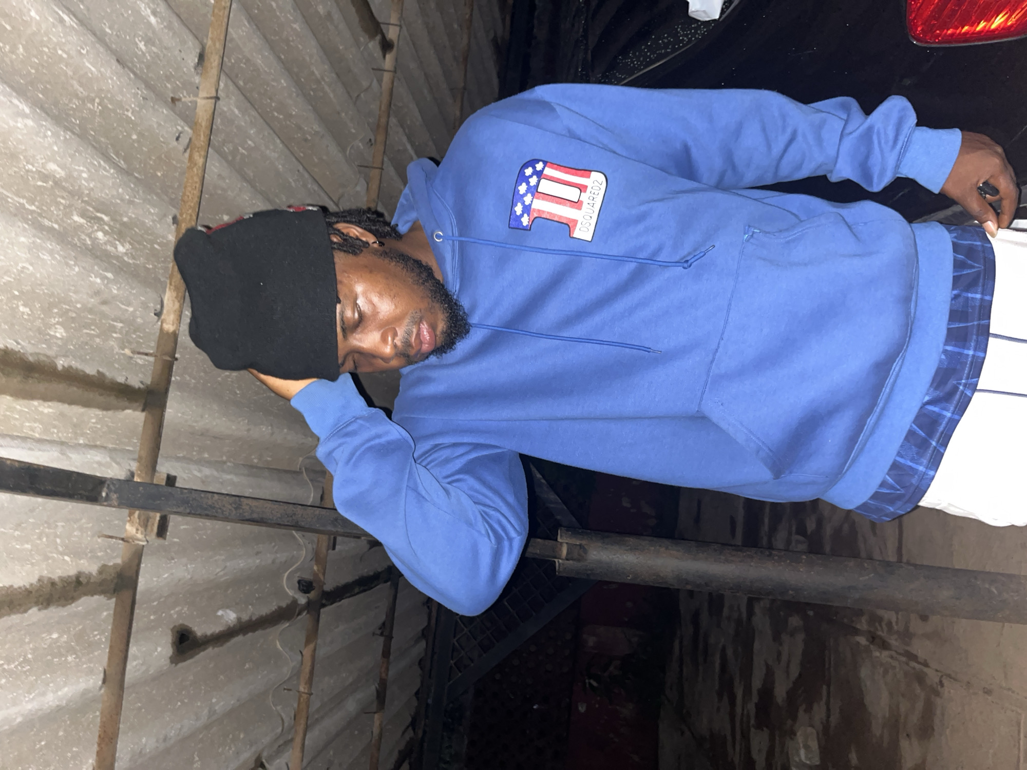 A full-shot photograph captures a Black male standing in a dimly lit outdoor area. He is wearing a blue hooded sweatshirt with a "DSQUARED2" logo on the left chest, black beanie, and white shorts with a blue plaid trim visible at the hem. His right hand is raised to his head, and he is looking down. In the background, corrugated metal roofing is supported by metal beams, and a black vehicle is parked to his right. The ground appears to be damp and littered. The image seems to be taken in Jos, Nigeria, based on contextual information.