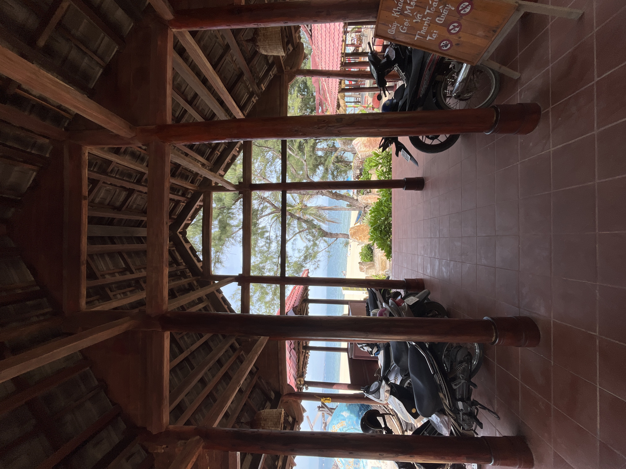 This is a photograph taken from within a covered wooden structure, looking out towards a beach scene in Mũi Né, Vietnam. The structure features a prominent wooden roof with exposed beams and multiple large wooden posts supporting it. In the foreground, several motorcycles are parked on a terracotta tiled floor. The view through the open sides of the structure reveals a sandy beach, the blue ocean, and lush green trees, including palm trees. Red-tiled roofs of smaller buildings are visible further down the beach. The sky is bright and appears to be a clear day. A wooden sign with Vietnamese writing and icons is partially visible on the right side of the frame. There are no people clearly visible in the immediate foreground or mid-ground.