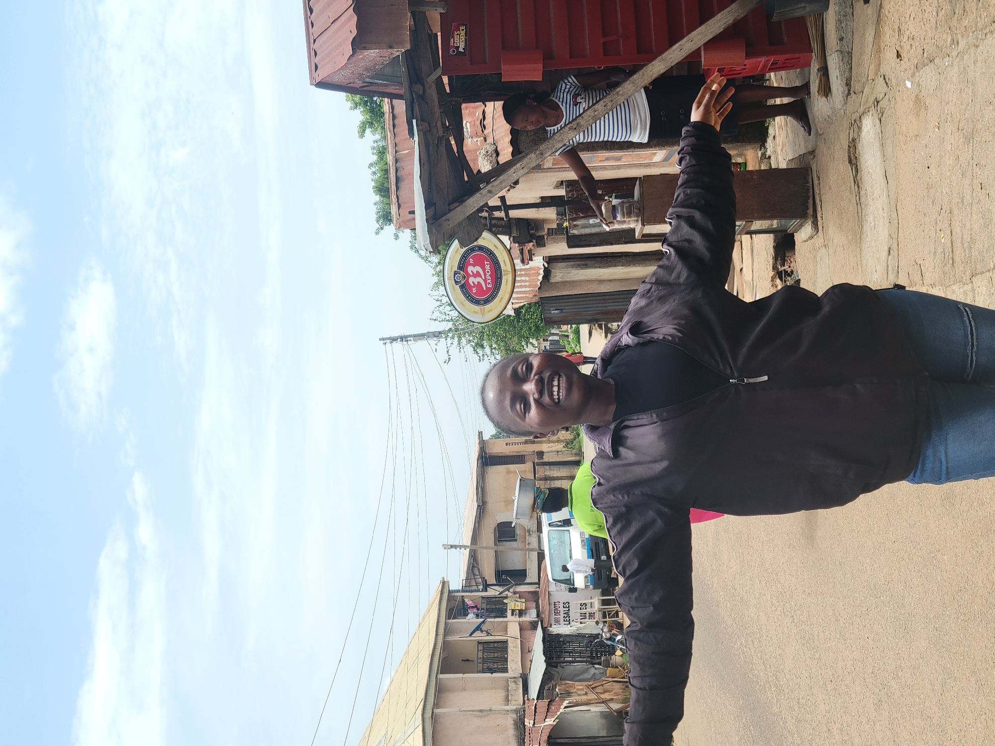 The image captures a street scene in Jos, Nigeria. A woman is prominently featured in the foreground, smiling and with her arms raised. She wears a dark jacket and blue jeans. Behind her, there are buildings, one with a sign that reads "GARRI DEPOTS LESALES." A white van is parked nearby. A "33" Export beer sign is visible. The road appears to be paved and somewhat worn. Another person is present, standing at a food shack. Overhead, there are power lines against a slightly cloudy sky.