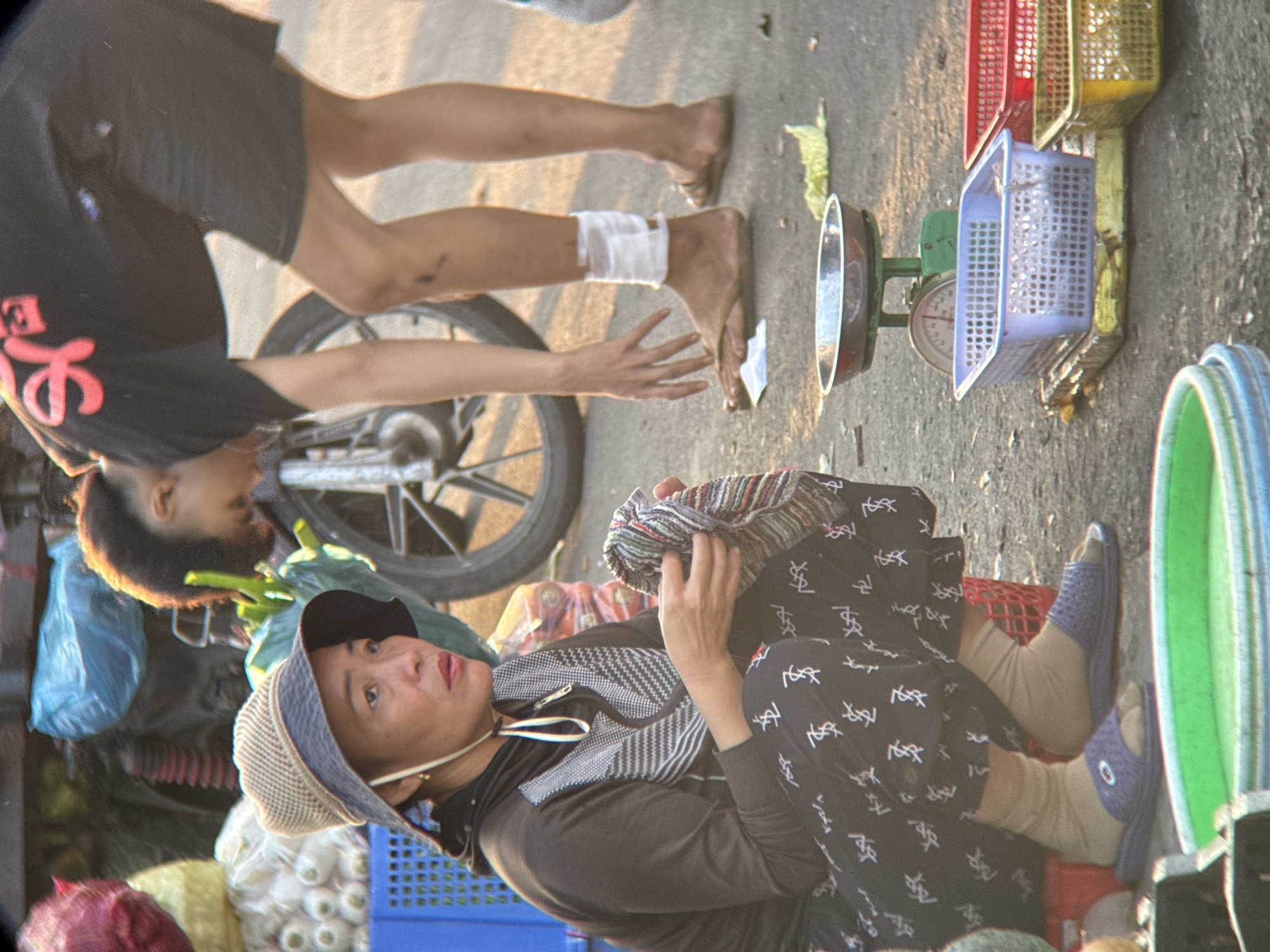 The image depicts a street scene in Nha Trang, Vietnam, likely a market. In the foreground, a woman wearing a patterned black outfit, a light-colored hat, and beige socks with blue sandals is kneeling and holding a striped cloth. Her expression is somber. To her right, a person in a black t-shirt and dark shorts is bending down, with one leg bandaged around the ankle. A motorcycle wheel and parts are visible behind them. The ground is asphalt, and various items are present: a green and silver scale with a metal bowl on top, blue and red plastic baskets, and some produce bags in the background. The lighting suggests it is either early morning or late afternoon.