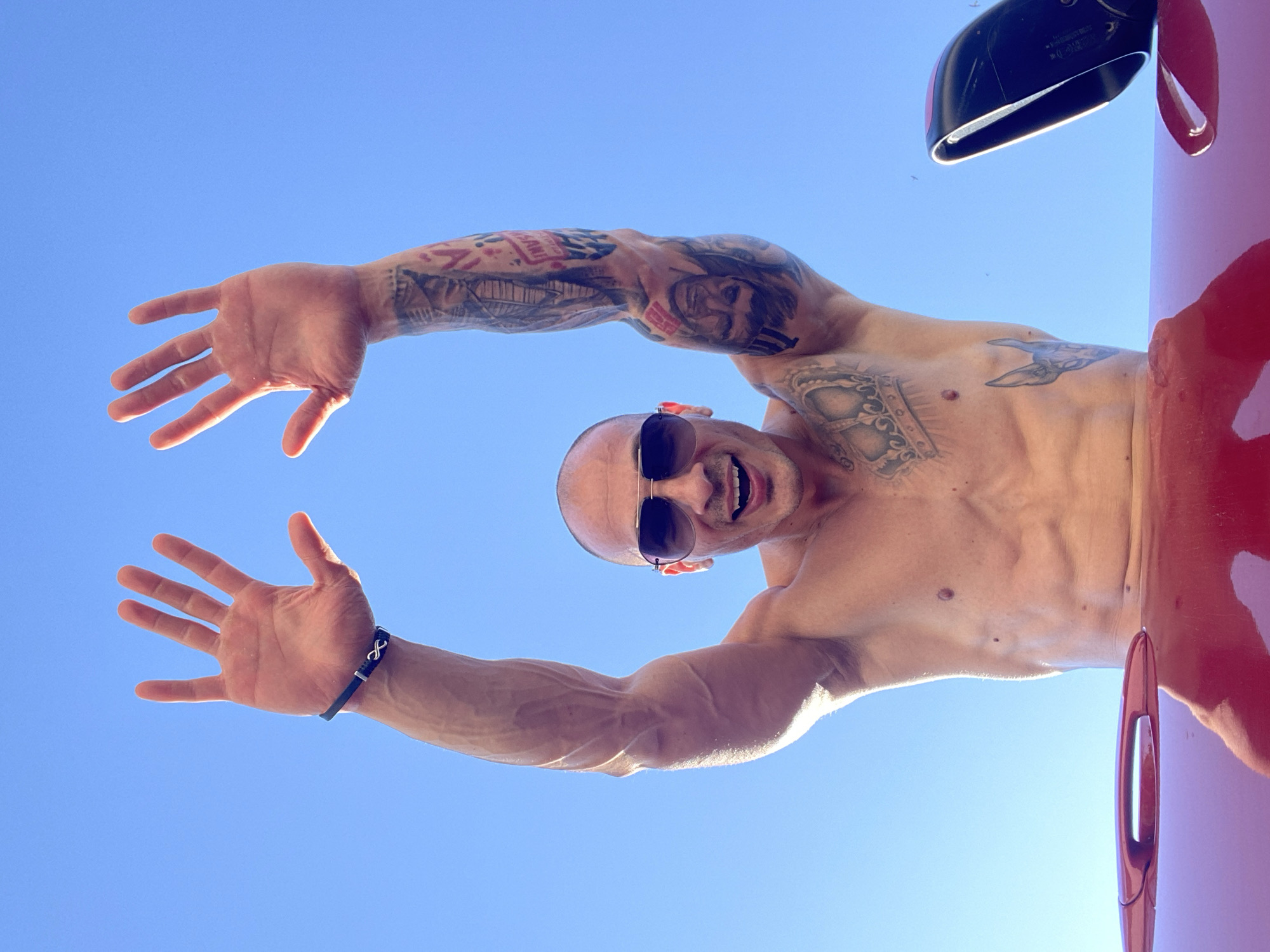 An unidentifiable shirtless man with tattoos on his arms and torso, wearing sunglasses and a bracelet, raises his hands towards a clear blue sky. He is leaning over the edge of a red vehicle, possibly a car, with its door handle and part of its body visible in the foreground. The image is taken from a low angle, looking up at the man against the sky.