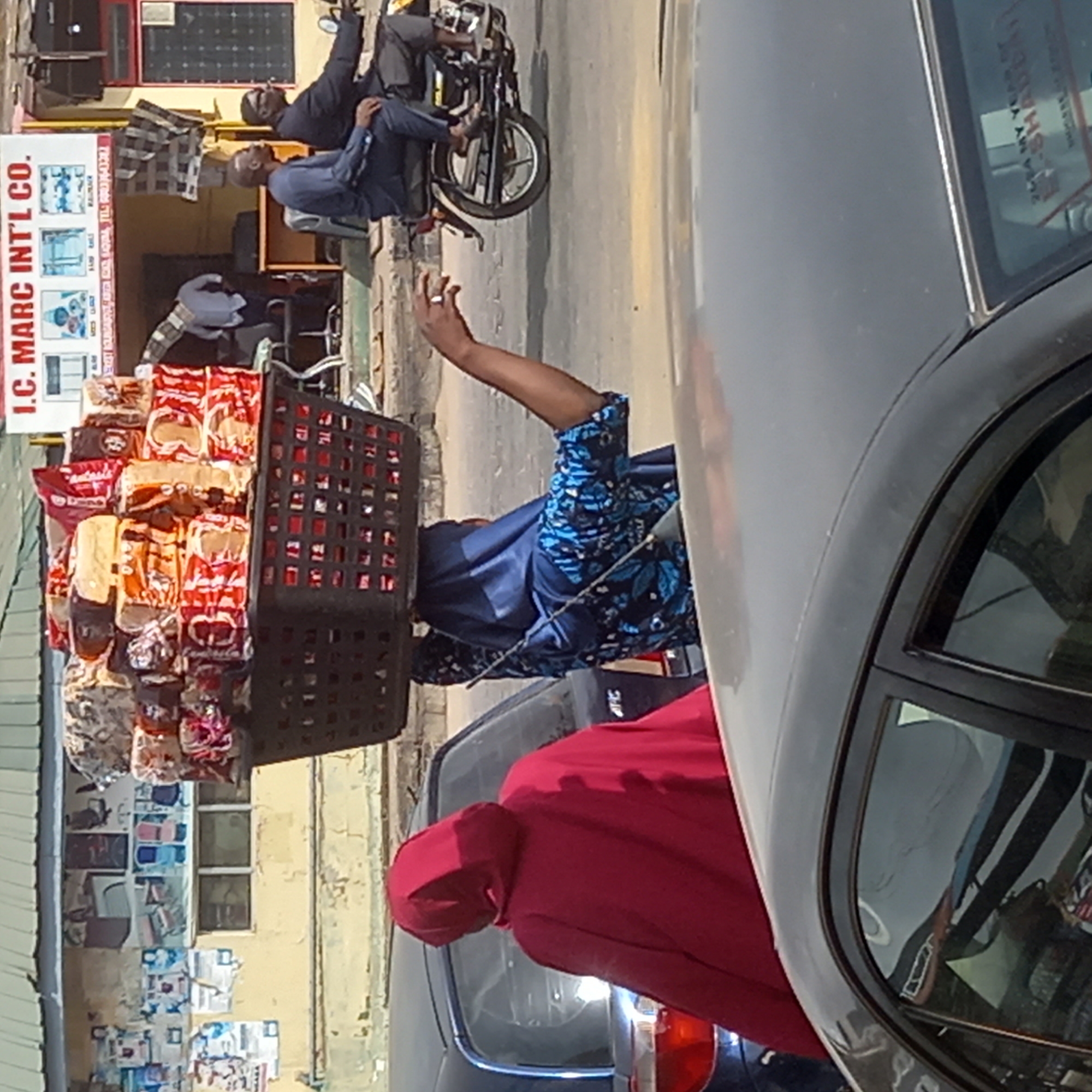 The image shows a street scene in Dadi Riba, Nigeria. A woman in a blue and floral patterned shirt and a dark undershirt carries a basket filled with packaged bread on her head. Her right arm is raised. She is positioned between two cars; one is a gray sedan on the right, and the other is a black vehicle with a red hood and tail lights on the left. In the background, there are two men on a motorcycle parked in front of a building, on top of which a solar panel is set, and an I.C. Marc Int'l Co. sign is visible. The building's wall is covered with posters.