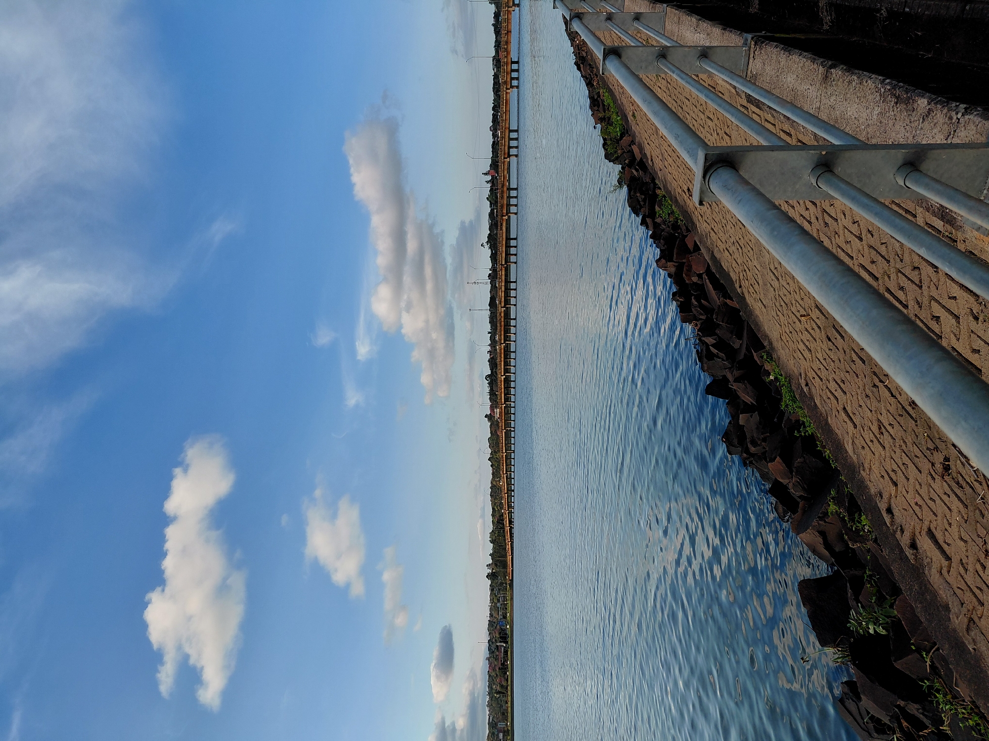 The image shows a wide shot of a body of water, likely a river or lake, in Encarnación, Paraguay. The water is calm with ripples reflecting the blue sky and scattered white clouds. The foreground features a paved walkway with a metal railing, bordered by large, dark rocks and some sparse greenery. In the mid-ground, a long bridge stretches across the water, connecting to a distant shoreline dotted with buildings and trees. The sky is a clear blue with wispy white clouds, suggesting a pleasant day. There are no people visible in the image. The scene appears to be a public waterfront area, possibly a park or promenade, offering a view of the bridge and the cityscape beyond.