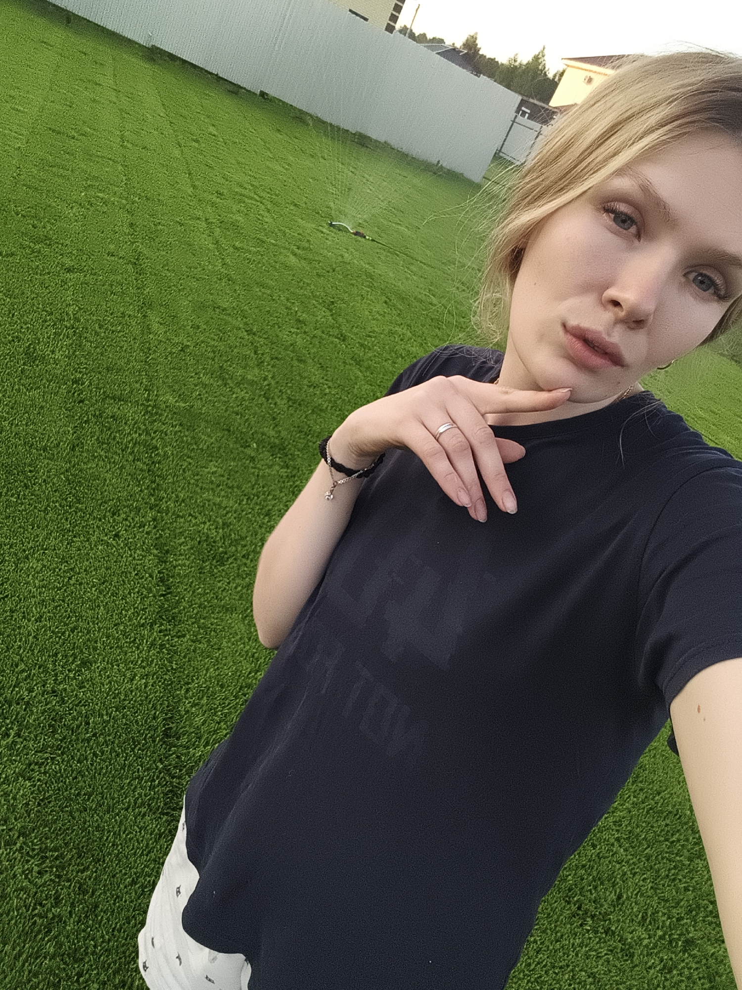 A young, light-skinned woman with blonde hair stands on a green, grassy surface, possibly artificial turf. She wears a dark-colored t-shirt and light-colored shorts. Her left hand is raised, with her fingers pointing towards her chin. She is wearing a ring on her left ring finger and a bracelet on her left wrist. The background features a white fence and a building in the distance. The image appears to be taken from a slightly elevated angle. There is no evidence to suggest this picture was taken in Русский Вожой, Russia.