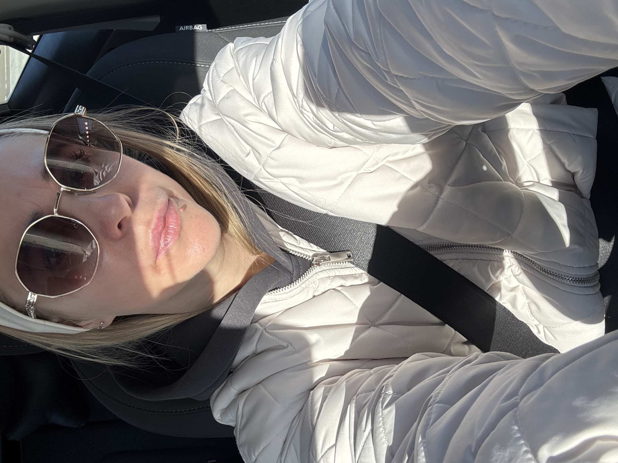 A woman with blonde hair is seated in the driver's seat of a vehicle, secured with a seatbelt. She is wearing a white quilted jacket, a gray hooded sweatshirt, and round sunglasses. A white headband is visible in her hair. The interior of the car includes black leather seats and a dashboard with an "AIRBAG" label. The image is taken in bright sunlight, creating shadows across the woman's face and jacket. There is no indication that this image was taken in Chaikovsky, Russia.