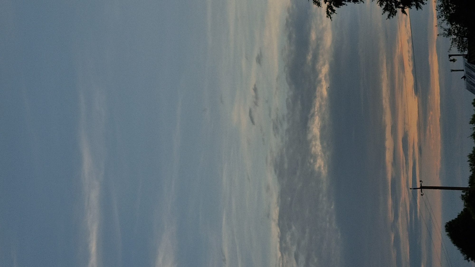 A vertical, eye-level, medium shot captures the sky above the city of Vysokoe, Russia. The sky is a soft blue, transitioning to pale orange near the horizon, and is filled with thin, wispy clouds in varying shades of gray and white. The lower portion of the image features the silhouettes of trees and a power line pole against the colorful sky. A dark tree branch extends into the frame from the right side, and the rooftop of a building with a chimney is visible at the bottom right. The scene appears to be set during dusk or dawn.