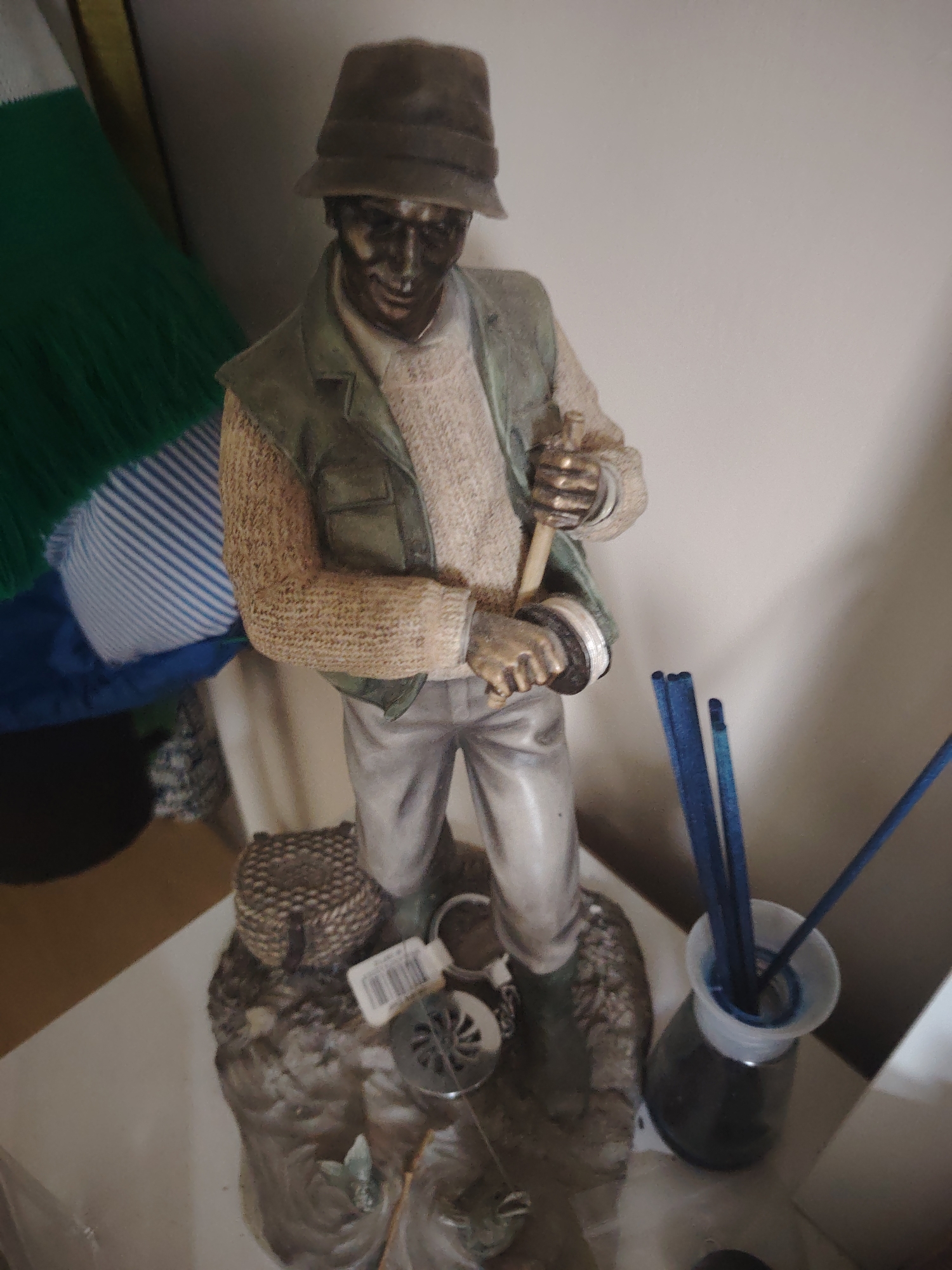 The image shows a statue of a fisherman standing on a base. The fisherman is wearing a hat, a vest over a sweater, and pants. He is holding what appears to be a fishing rod. In the background, there is a green and blue striped fabric, and a wall. To the right of the statue, there is a glass bottle with blue reeds inside, possibly an air freshener. The statue is placed on a white surface. There is no indication of the city of Huntingdonshire, United Kingdom in the image.