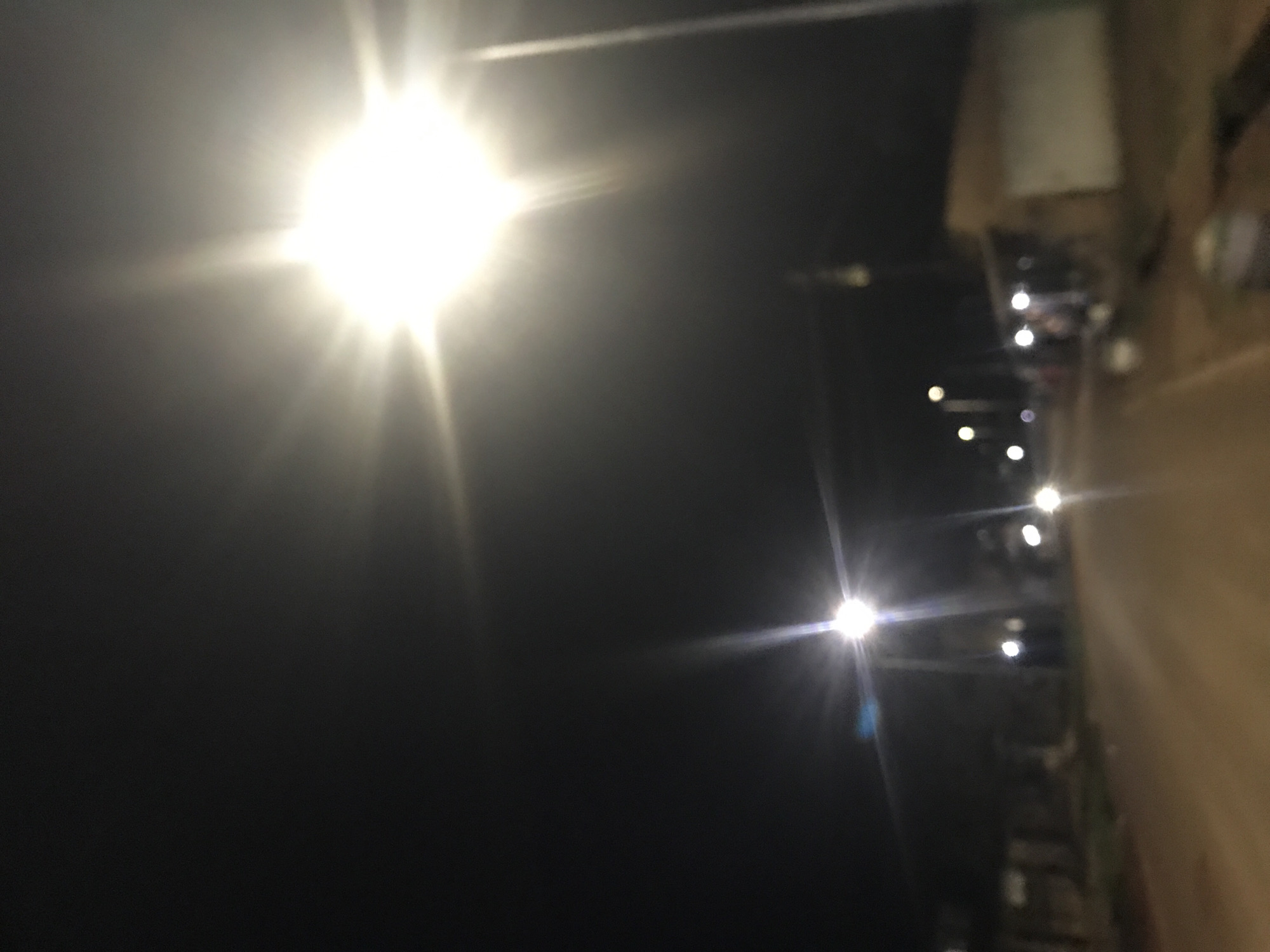 The image captures a dimly lit street scene at night in the city of New Stadium, Nigeria. The primary focus is a bright street lamp in the upper right quadrant, casting a strong glare that obscures details in that area. To the left and further down the street, other streetlights are visible as blurry orbs. The road appears to be unpaved, with a light brown surface. Buildings line the right side of the street, one of which has a light-colored wall. Silhouettes of people can be vaguely discerned near the buildings. The overall impression is a somewhat hazy, night-time urban environment.
