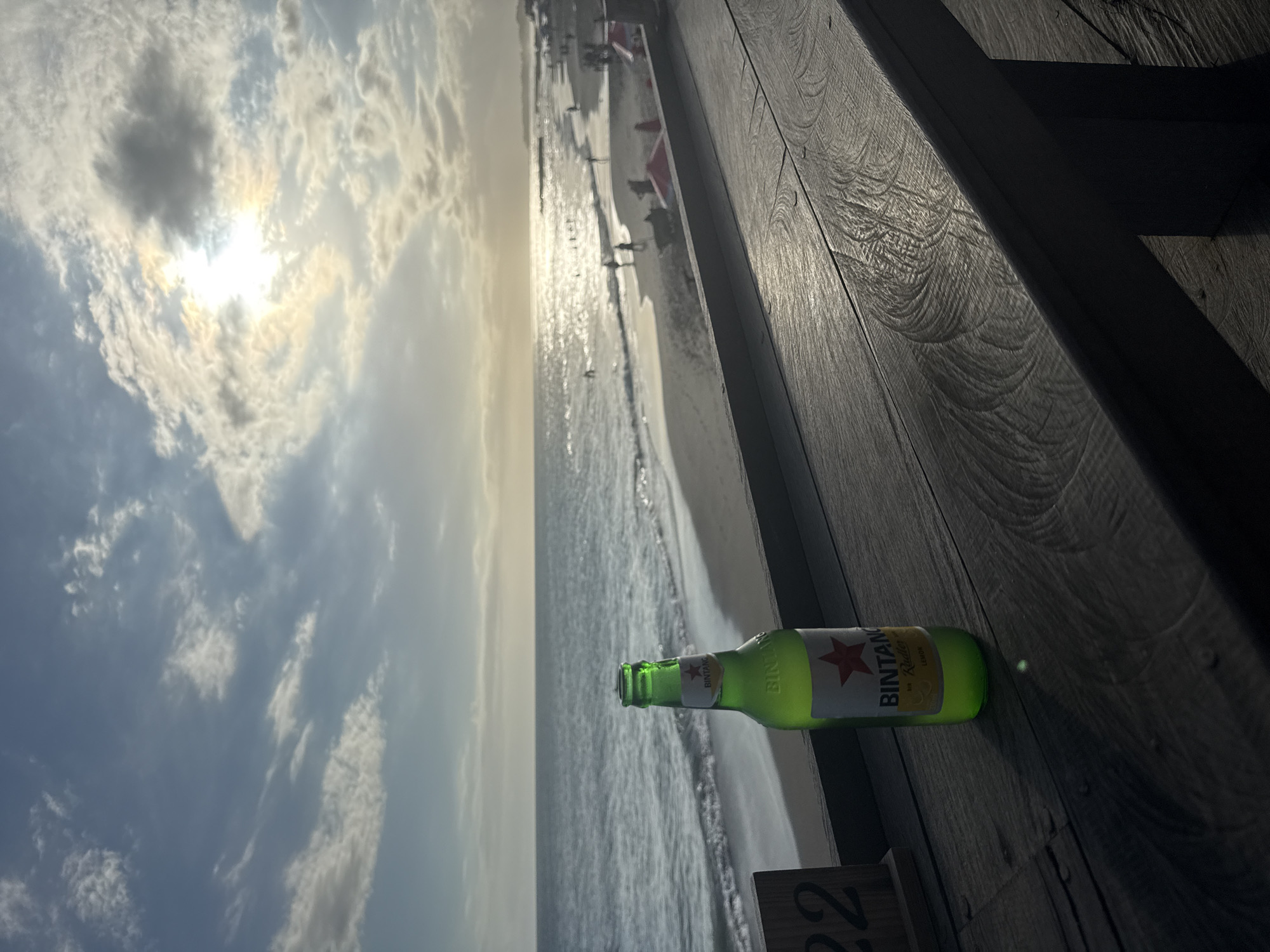 A horizontal shot captures a Bintang Radler beer bottle lying on a wooden deck, against a backdrop of a beach scene. The beer bottle, mostly green with a white label marked with a red star and the word "BINTANG", is positioned near the right edge of the frame. The wooden deck's planks run diagonally, occupying the top-right quadrant of the image. Beyond the deck, a sandy beach stretches along the shoreline, with faint figures of people visible in the distance. The ocean reflects sunlight, creating a glistening effect on the water's surface. Above, the sky is filled with clouds, partially obscuring the sun. The location is identified as North Kuta, Indonesia.
