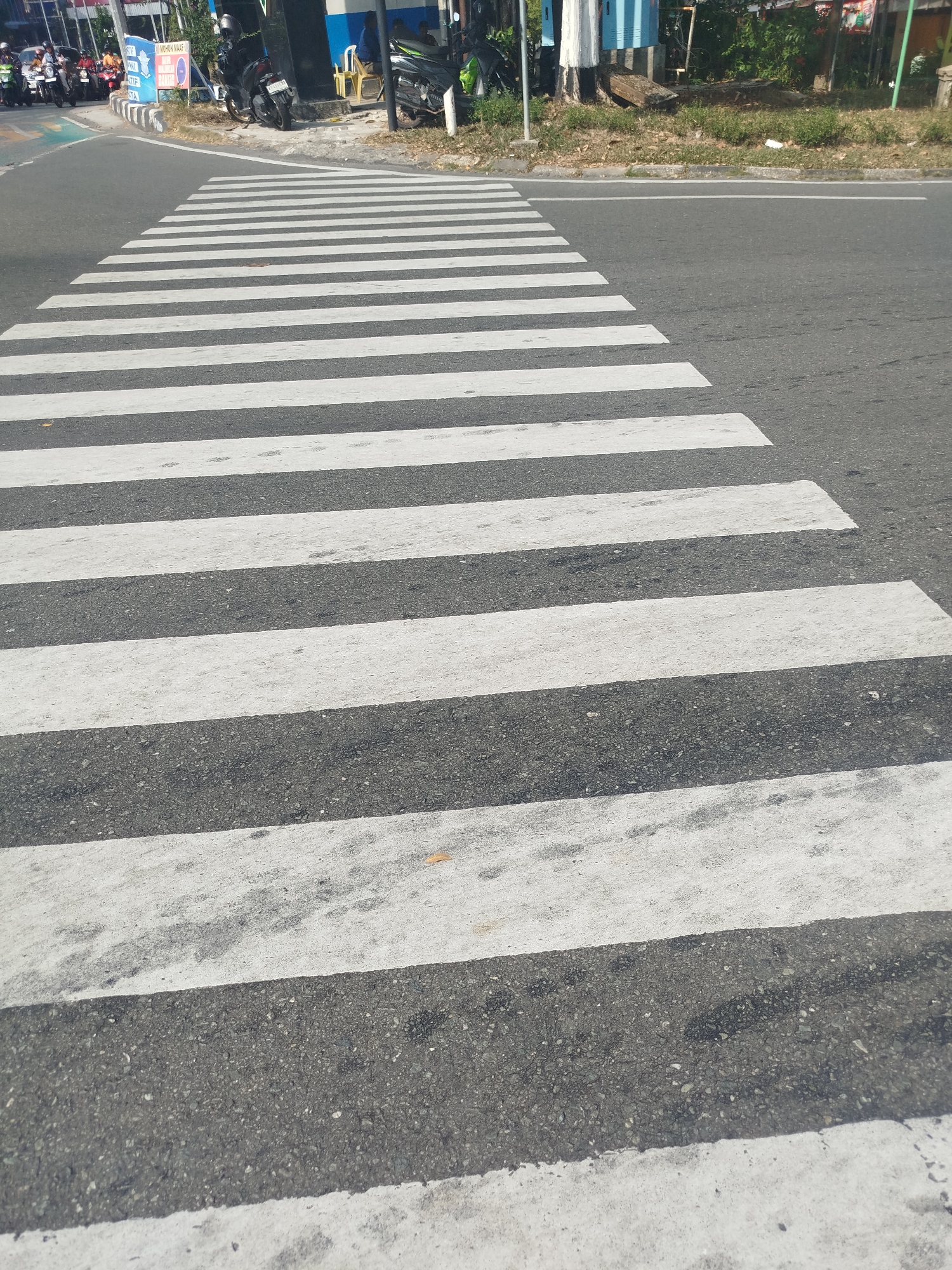 The image captures a perspective shot of a pedestrian crossing on an asphalt road in Balikpapan, Indonesia. The crossing is marked with alternating white and dark gray stripes, indicative of a standard zebra crossing. In the background, a sidewalk area is visible, bordered by greenery and some structures. To the left of the pedestrian crossing, there is a traffic island with signage and several motorcycles parked. A few individuals are seen near the motorcycles and on the sidewalk. The road surface appears worn in places, with visible aggregate in the asphalt and some fading on the white stripes of the pedestrian crossing. The lighting suggests it is daytime, with shadows cast on the road.