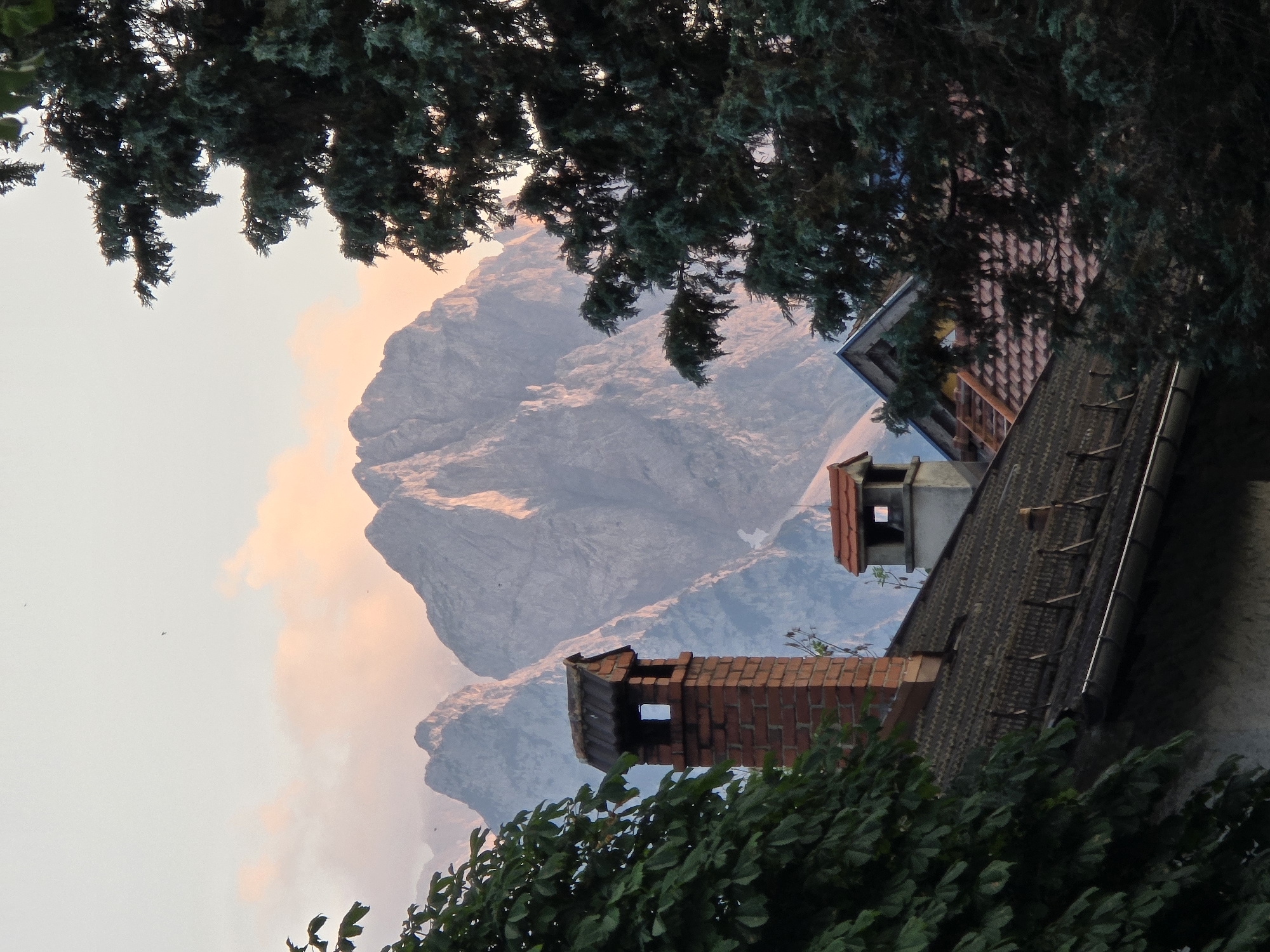 This is a photograph taken in Garmisch-Partenkirchen, Germany, focusing on a mountain landscape behind a residential area. The scene captures a view of imposing, jagged mountains in the background, bathed in the warm light of either sunrise or sunset, with pink and orange hues coloring the sky and the peaks. In the foreground, several rooftops of houses are visible, constructed with dark tiles. Prominent chimneys stand on these roofs; one is made of red brick with a square opening, and another is smaller and made of stone. Dense green foliage from trees frames the left and right sides of the image, partially obscuring the view of the houses and the mountains. The overall impression is a juxtaposition of natural grandeur and human habitation.