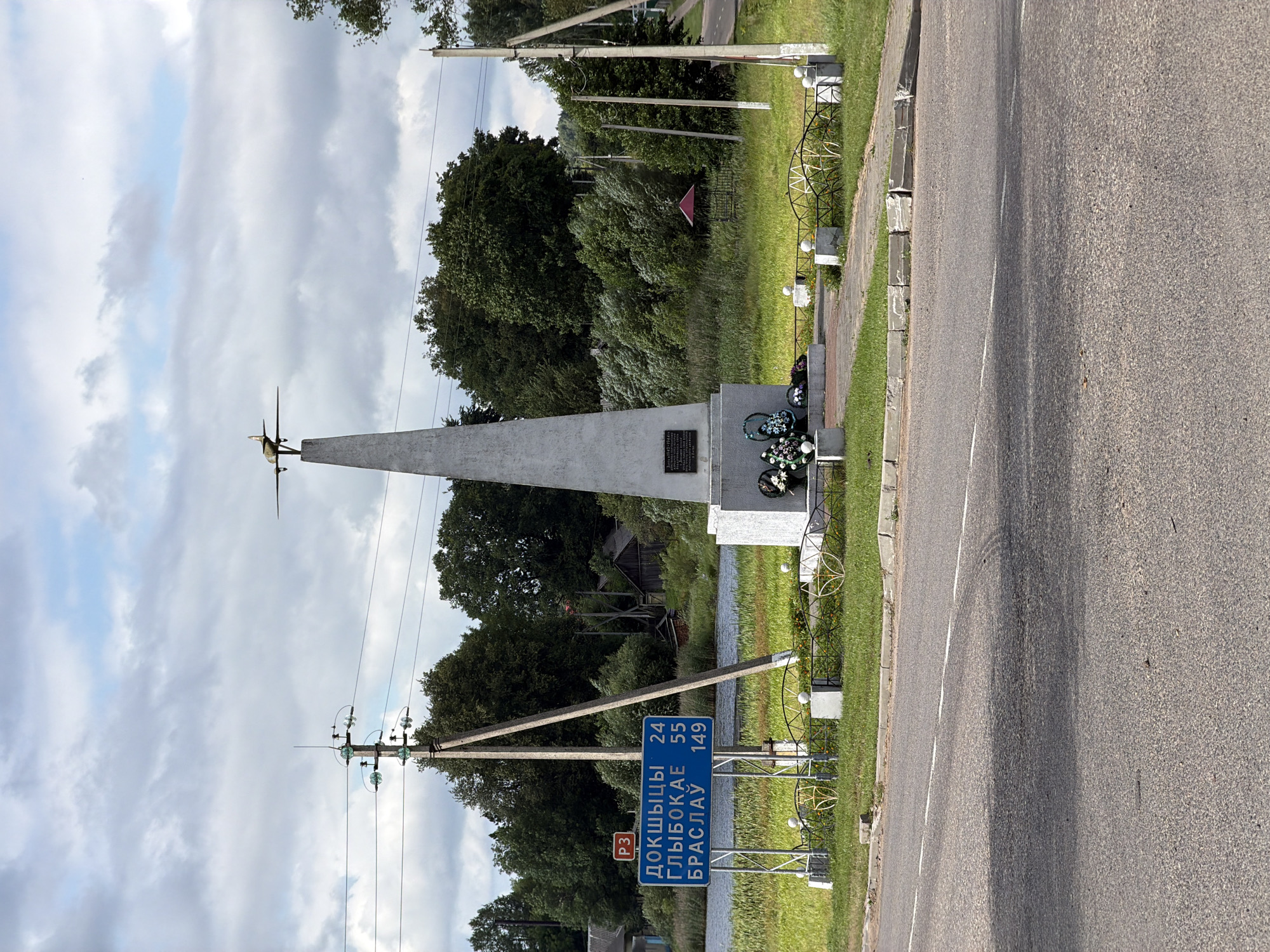 This media file captures an outdoor scene with a monument, road, and sky. The monument is a tall, slender, light-colored obelisk with what appears to be a model airplane or propeller on top. At the base of the monument, there's a plaque and a wreath of artificial flowers. A decorative metal fence surrounds a portion of the monument and adjacent grassy area.
The scene includes a paved road running alongside the monument, with a distinct white line marking its center. There's a grass verge and a sidewalk between the road and the monument. Utility poles with electrical wires are visible, one of which is near the monument and another in the background.
In the background, there are dense trees and some vegetation. A small red-roofed structure is partially visible amongst the trees, suggesting a rural or suburban setting. The sky is partly cloudy, with a mix of light and darker gray clouds, indicating it could be daytime with variable weather.
A prominent blue road sign is mounted on a pole near the monument. It displays Cyrillic text and numbers: "ДОКШЫЦЫ 24", "ГЛЫБОКАЕ 55", and "БРАСЛАУ 149". This sign indicates distances to the towns of Dokshytsy, Hlybokaye, and Braslav, suggesting the location is on a road connecting these places. There's also a red square sign with "РЗ" inside, which is likely a road designation.
There are no people visible in the frame. The overall impression is a roadside memorial or landmark in a natural setting. The lighting suggests it's daytime, likely late morning or afternoon given the shadows. The presence of the monument and the road sign point to this being a significant location along a travel route.