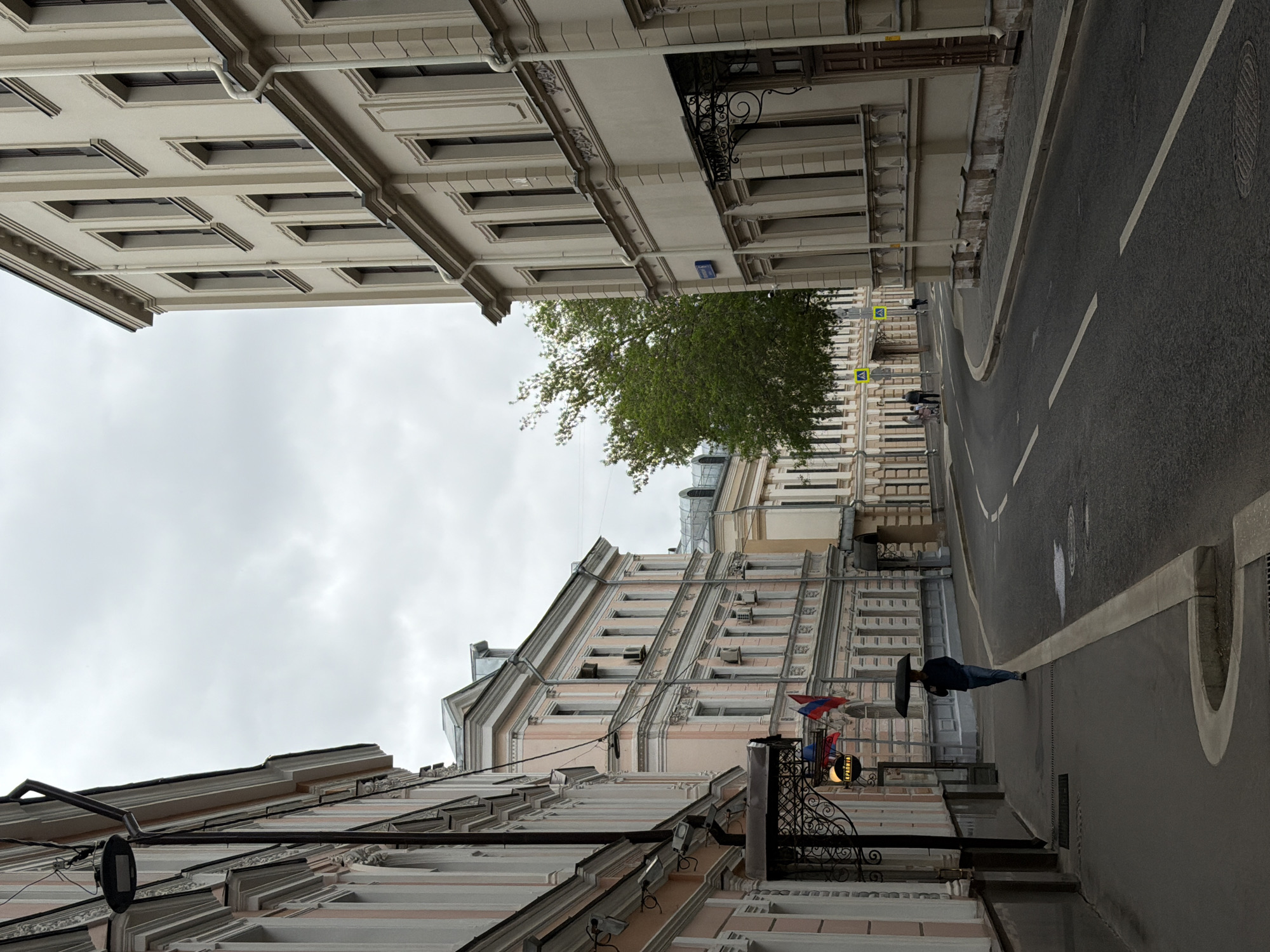 The image depicts a street scene in Moscow, Russia, shot at an unusual angle. The buildings on either side of the street are tall, with ornate facades and multiple windows. The building on the left is pink with decorative architectural details. The building on the right is white and has a balcony with ironwork. A green tree is visible between the buildings. The street is paved and has a white line marking the lane. A person is walking on the sidewalk with an umbrella. The sky is overcast.