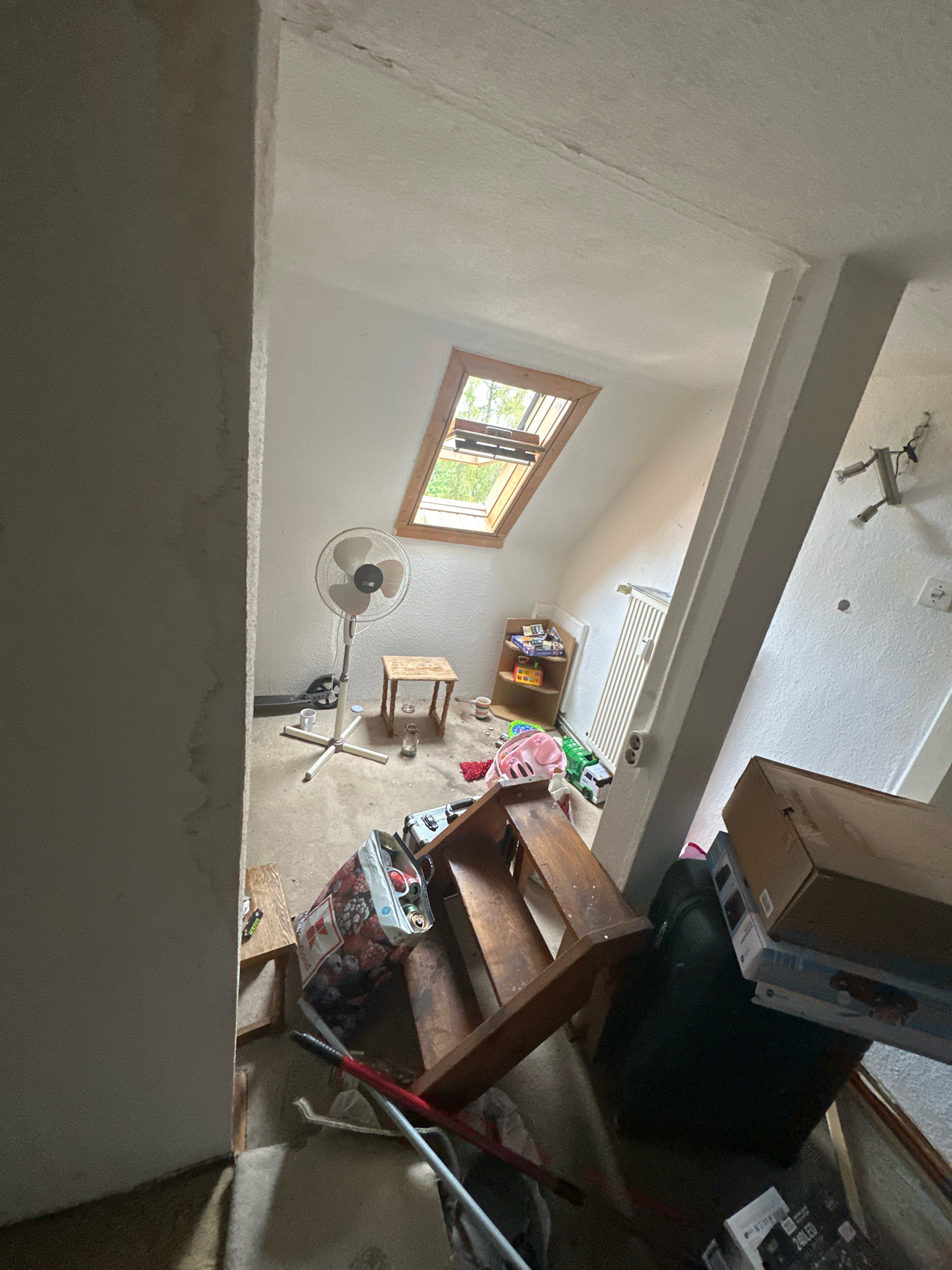 This is a wide-angle, medium-shot, eye-level interior photograph of a cluttered attic room. The room appears to be a storage space or a disused living area. The walls are painted white and appear textured. A pitched ceiling slopes from right to left, with a wooden-framed skylight at the apex. A vertical support beam, also painted white, bisects the right side of the image.
In the foreground, a wooden step stool or small bench is overturned, with a colorful fabric bag containing cans and other items placed on it. Various tools, including what appear to be cleaning implements, are scattered on the floor. Several cardboard boxes, some stacked and some open, are visible on the right side of the image, along with a large dark-colored suitcase.
Further into the room, a white oscillating fan stands on a white base. A small, rustic wooden stool is positioned behind the fan. On the floor are several glasses and mugs, and a red rug. A child's scooter is visible near the fan. In the background, to the left, a multi-tiered wooden shelf unit holds various toys and items, including a green toy truck and a pink object that may be a child's toy. A white radiator is mounted on the wall behind the shelf unit.
The overall impression is one of disarray and clutter. The lighting appears to be natural light from the skylight and possibly artificial light from an unseen source. The floor is concrete or a similar utilitarian material. No people are visible in the scene. The location of Bitterfeld-Wolfen, Germany, is not visually discernible from the image itself; it is provided as context.