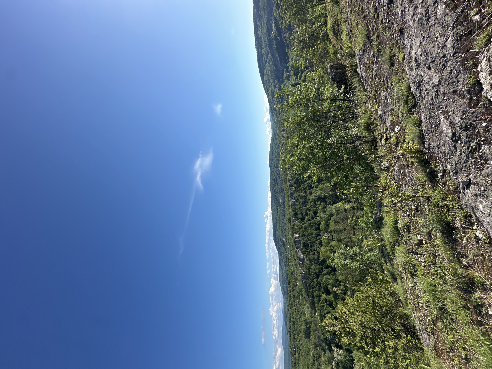 The image shows a landscape with a deep blue sky with a few wispy clouds. A mountain range covered in dense green forest runs along the right side of the image, with a rocky terrain in the foreground. The perspective suggests the photo was taken from a high vantage point, possibly on or near a cliff. Given the context of Lovech, Bulgaria, this could be a view from the Hisarya Fortress overlooking the city and surrounding valleys. There are no people or interactions visible in the image. The environment is characterized by a sunny, clear day and lush vegetation.