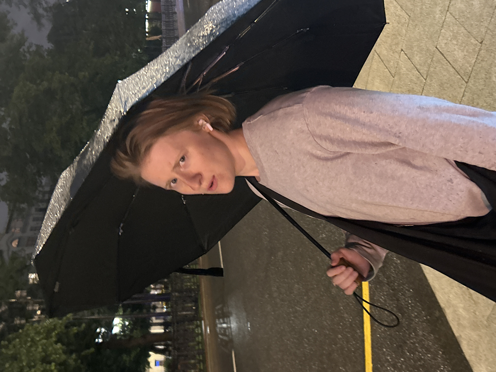 A person with blond hair and white earbuds is holding a black umbrella and a black bag while standing on a wet sidewalk at night in an urban environment. The person is wearing a light-colored sweatshirt. The umbrella's edge is decorated with a sparkly, reflective pattern. In the background, trees, buildings with lights, and a street with a yellow line are visible. The ground appears wet, suggesting it has been raining.