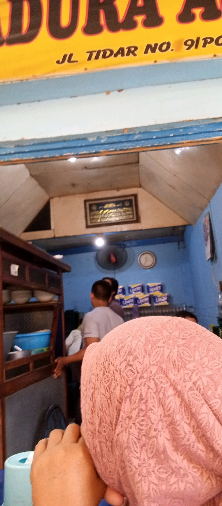 In a dimly lit interior, a person wearing a pink headscarf is partially visible in the foreground. The headscarf has a floral pattern. To the left, a wooden cabinet with shelves containing bowls is seen. Behind the cabinet, a blue wall is visible. In the background, a fan is mounted on the wall, and a clock is positioned above it. Stacks of what appear to be packaged goods, possibly beverages, are also present on a shelf against the blue wall. The environment suggests a food establishment or a shop. A yellow sign with black lettering is visible at the top, with the text "JL. TIDAR NO. 91 PO" clearly legible, indicating the location is likely on Jalan Tidar, Surabaya, Indonesia.