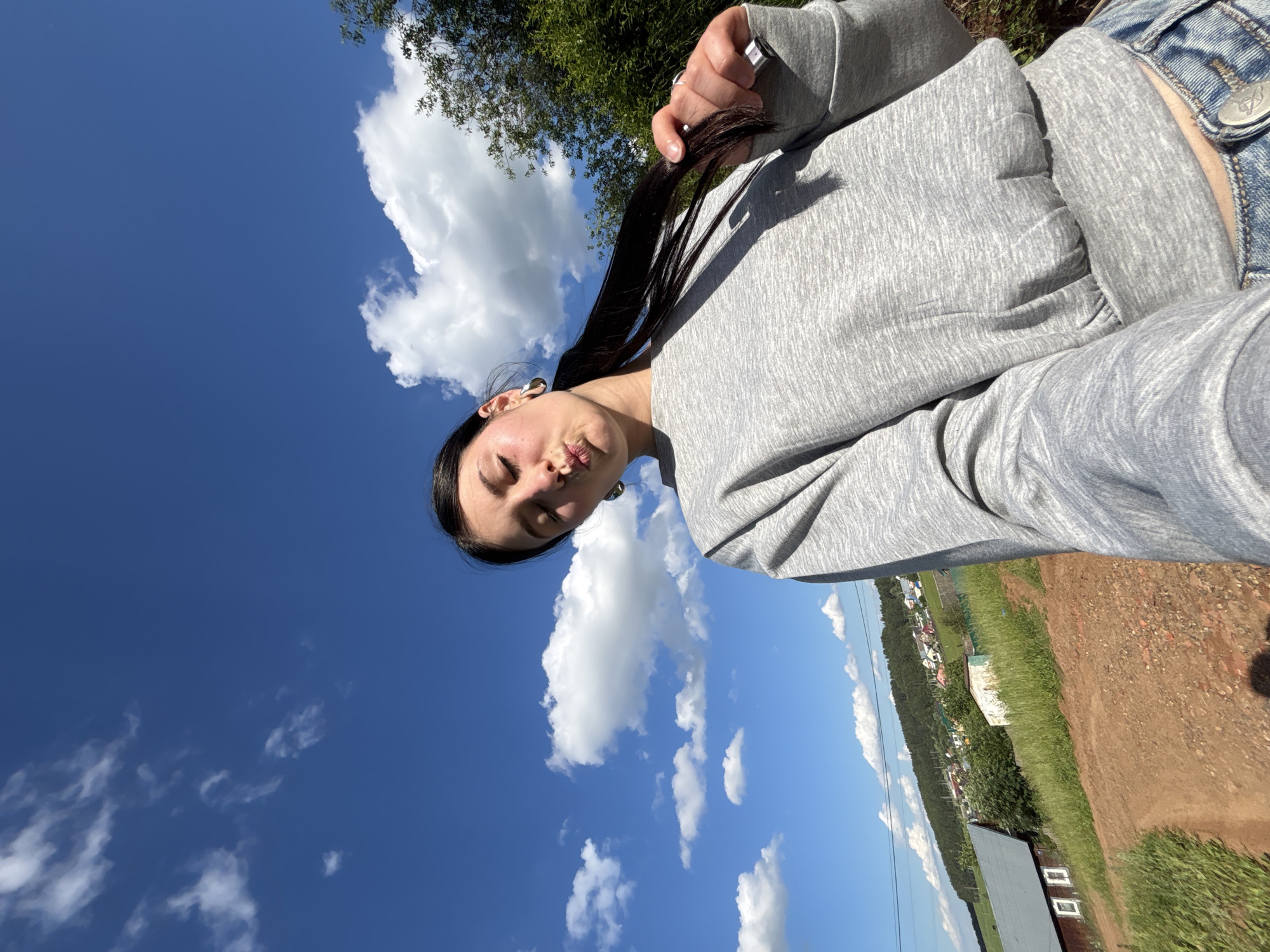 A young woman with dark hair is taking a selfie outdoors under a bright blue sky with fluffy white clouds. She is wearing a grey sweatshirt and blue jeans, and her eyes are closed as she puckers her lips. Her right hand is holding a section of her hair. In the background, there is a green, hilly landscape with a small village visible on the slope, featuring houses with red roofs and some trees. A dirt path leads up the hill in the foreground, with a wooden building to the left. The scene appears to be in a rural or semi-rural area, likely the village of Fokino, Russia, given the context. The lighting suggests it is daytime, likely late morning or afternoon, given the sunlight and shadows.
