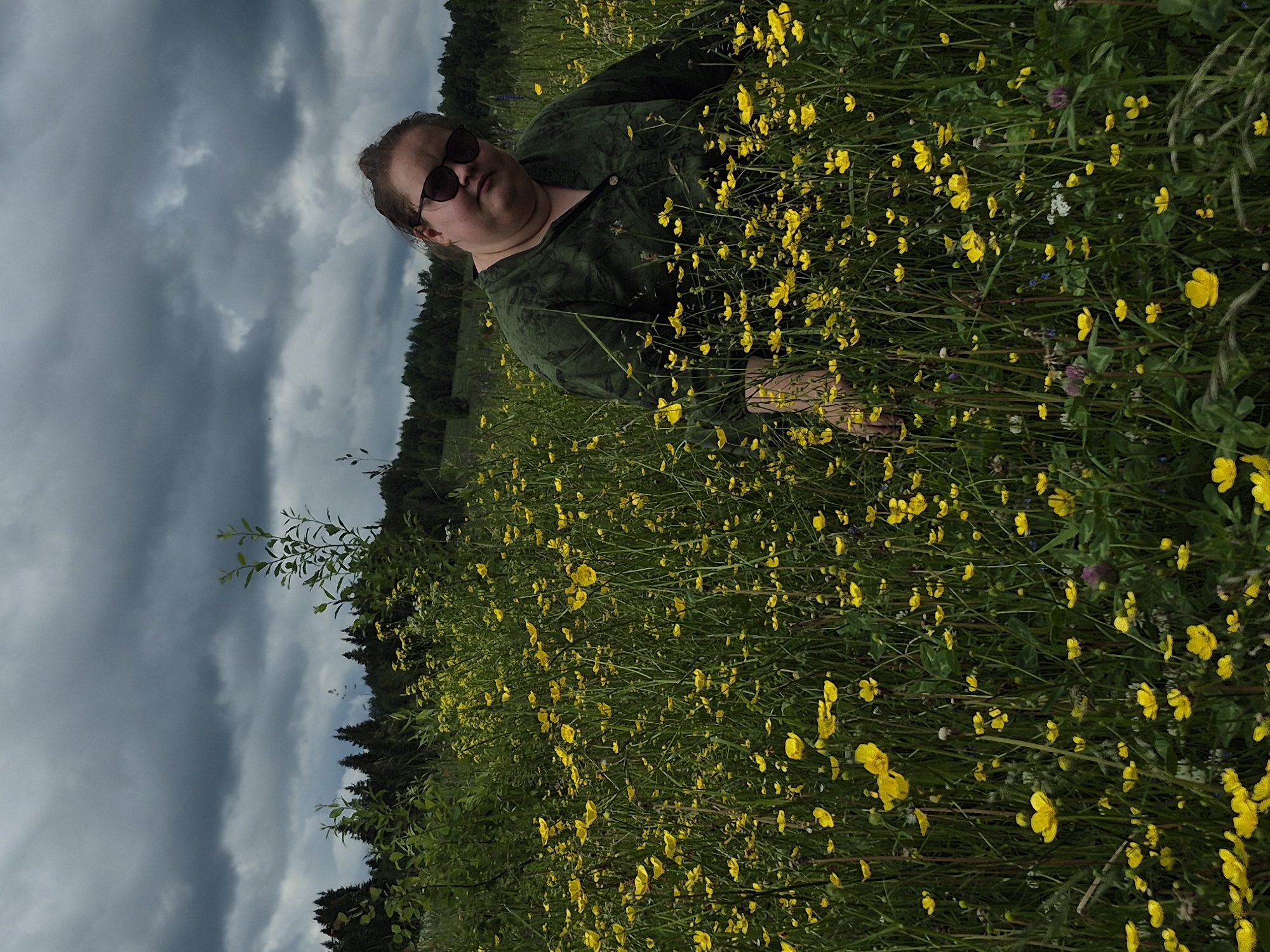 A person wearing sunglasses and a dark green, patterned shirt sits in a field of yellow wildflowers under a cloudy sky. The person is positioned in the lower right quadrant of the image, looking towards the viewer with a slight smile. The field is dense with tall green grass and numerous yellow buttercup-like flowers, with a dark forest line visible in the background. The location is described as a meadow in the vicinity of Dojnaya, Russia.