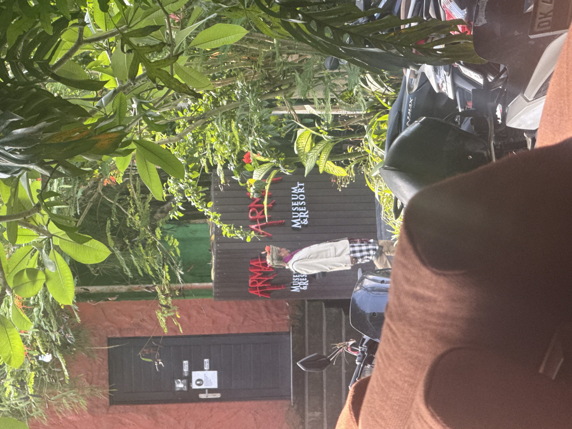 The image, rotated 90 degrees counter-clockwise, depicts an outdoor scene at what appears to be the entrance or parking area of a museum and resort, consistent with the Ubud District, Indonesia location.
In the foreground, a brown fabric object, possibly part of a bag or seating, partially obscures the view on the right. Directly behind it are several parked scooters or motorcycles, with a black helmet resting on one. A silver and black scooter, resembling a Yamaha NMAX, is clearly visible, along with another bike showing a partial Indonesian license plate "DK 4...".
The main subject is a rustic brown sign with red lettering that reads "ARMA MUSEUM & RESORT" (or "ARMA MUSEUM & RES"). Standing in front of this sign is a statue of a person, possibly a traditional Balinese figure, wearing a white jacket, a checkered sarong, and a hat or head covering.
To the left of the sign, an orange or red-hued building with a dark wooden or metal door is visible. There are steps leading up towards the area with the sign. The entire scene is surrounded by abundant, lush tropical vegetation, including large green leaves, hanging vines, and smaller bushes, indicating a verdant, natural setting.
The lighting suggests it is daytime, likely mid-morning or afternoon, with bright sunshine filtering through the dense foliage, creating dappled light and shadows. No active event is taking place; it's a static depiction of the location's exterior.