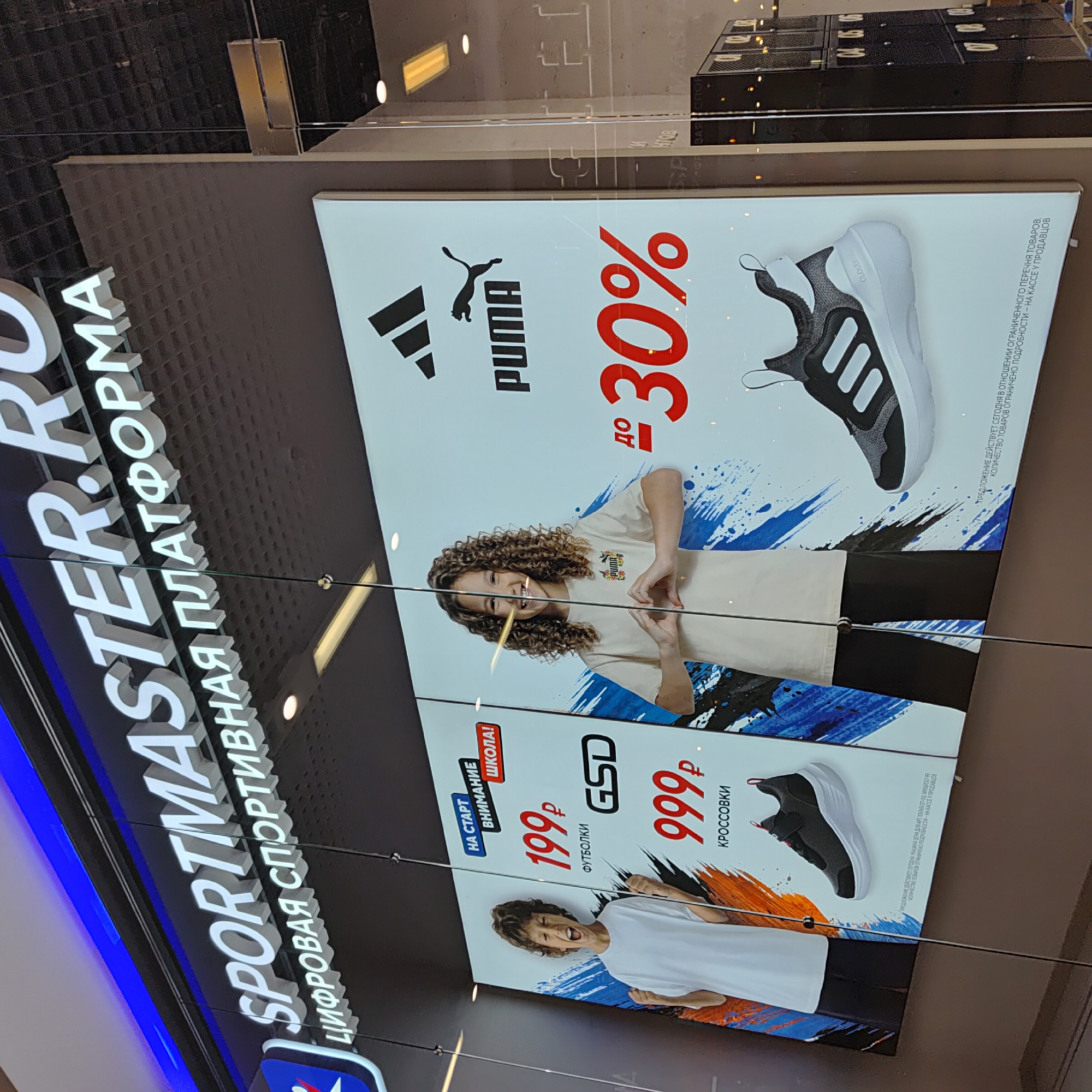 The image shows a storefront display for "SPORTMASTER.RU," a digital sports platform in Ufa, Russia. The display features promotions for t-shirts priced at 199 rubles and sneakers priced at 999 rubles, with a discount of up to 30% on some items. A boy wearing a white t-shirt and black pants is depicted on the left, and a girl with curly hair wearing a beige t-shirt is on the right. Prominent brand logos for Adidas and Puma are visible. The storefront is illuminated and appears to be in an indoor shopping environment, possibly a mall, as indicated by the ceiling lights and reflective surfaces. The display utilizes bright graphics and large font sizes to attract attention.