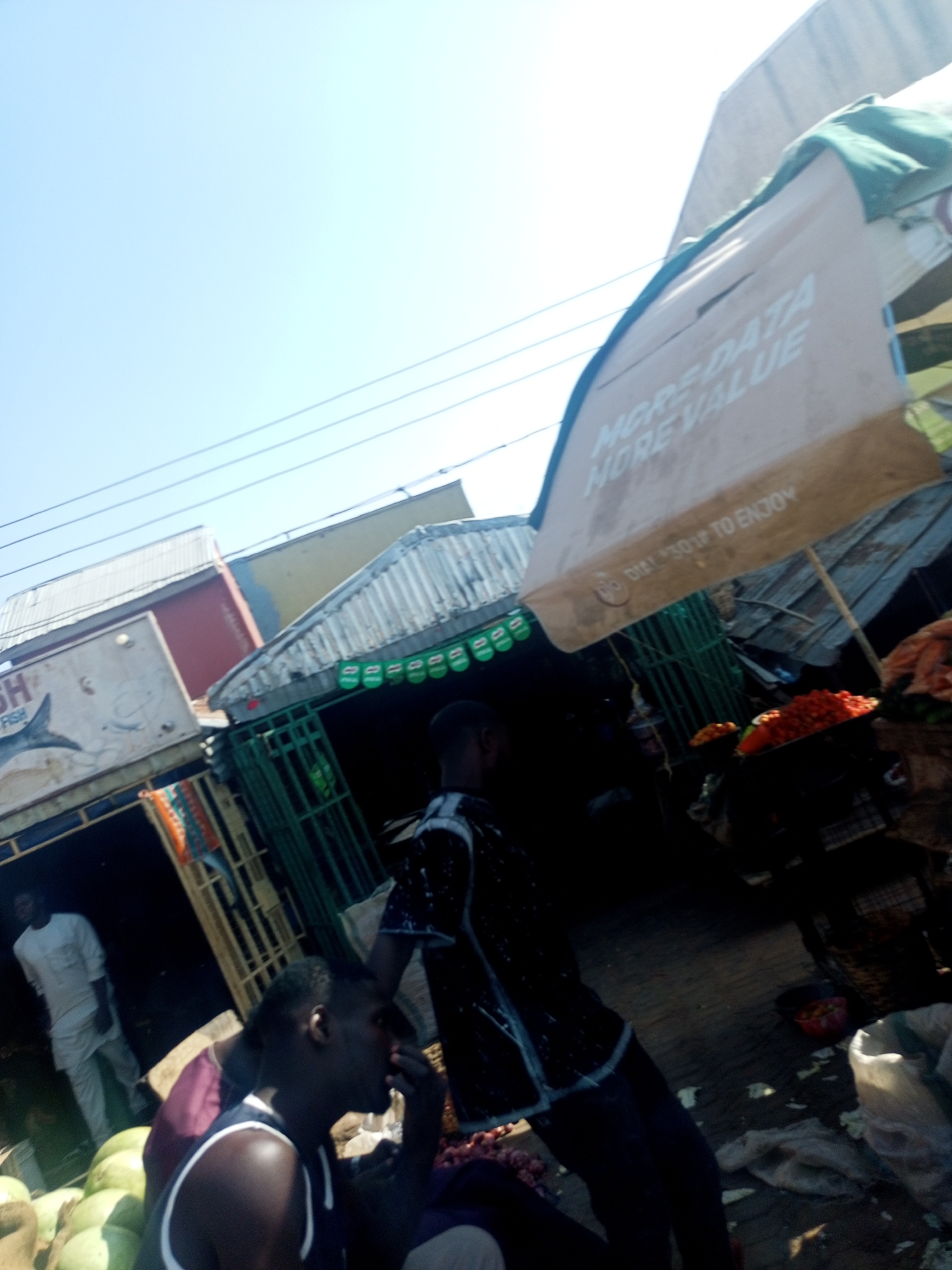 This image depicts a bustling outdoor market scene in Gombe, Nigeria, captured on a bright, clear day with a noticeable tilted perspective.
In the foreground, a man with dark skin, wearing a dark tank top with white trim, is seated or crouched, facing towards the right side of the frame with his hand near his face. To his left, several large green fruits, likely watermelons, are piled on the ground. Another man in a dark, patterned shirt stands further back on the right, partially obscured. On the far left, a man in a white or light-colored garment stands within the entrance of a market stall.
The market is characterized by several simple structures with corrugated iron roofs. On the left, a stall prominently displays a sign that clearly reads "SH" and "FISH," accompanied by a graphic of a fish. Towards the center-right, another stall features green metal bars or gating. A large, light-colored advertising umbrella or awning extends from the right, partially shading the market area. Text visible on this umbrella advertises "MORE DATA MORE VALUE" with the phrase "TO ENJOY" below it, though partially obscured. Underneath the umbrella, vibrant piles of red vegetables, likely tomatoes or peppers, are arranged on a stall for sale. The ground is visible with scattered sacks and debris, typical of a busy market environment. Thin electrical wires traverse the expansive, light blue sky above the market. The scene captures the essence of daily commerce and interaction within a local market setting.