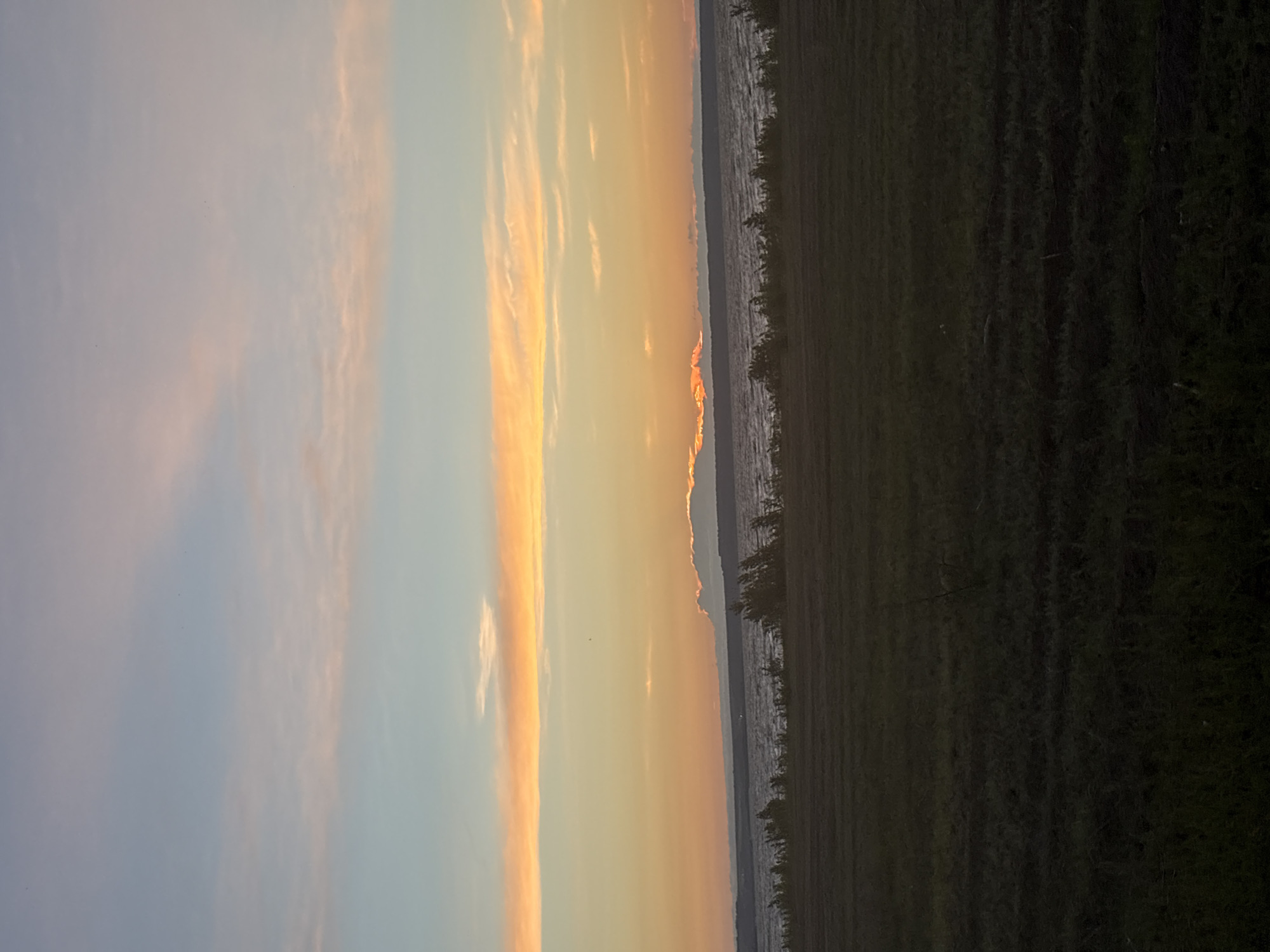 The image depicts a wide landscape scene at sunset. In the foreground, a dark, cultivated field stretches across the lower portion of the frame. Beyond the field, a body of water, likely a river or lake, occupies the middle ground. The water appears choppy, with visible waves. A line of low-lying vegetation and trees runs along the far bank of the water. The sky is a prominent feature, displaying a gradient of colors characteristic of sunset. Soft, horizontal bands of pale pink and orange clouds are visible against a fading blue sky. The sun itself is not directly visible, but its presence is indicated by the warm illumination of the clouds and the horizon. There are no people or specific buildings visible in the image. The scene appears to be in a rural or semi-rural setting. The location is identified as Alexeyevskoye, Russia.
