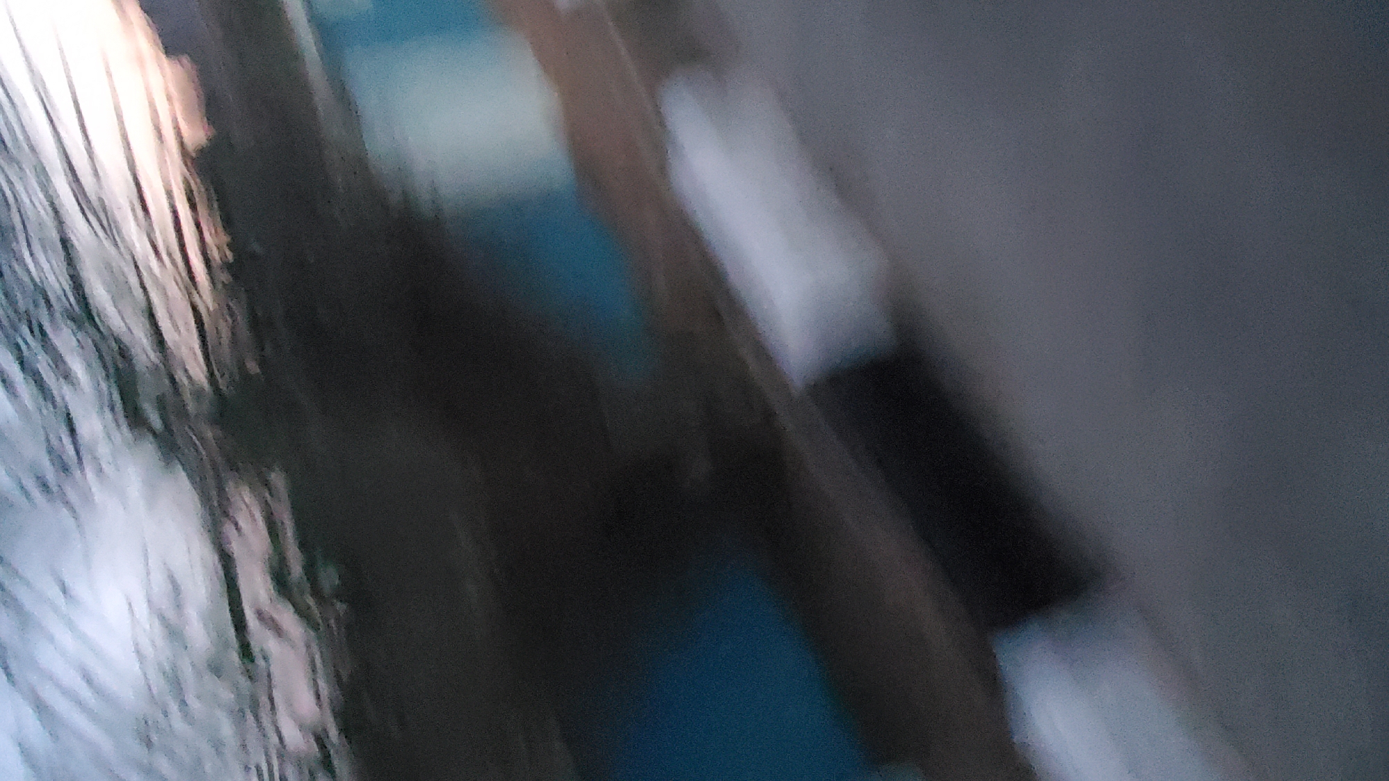 This image, captured in Jalingo, Nigeria, depicts an outdoor scene characterized by significant motion blur, indicative of being photographed from a moving vehicle. The upper portion features a streaked blend of light and dark grey tones, representing a blurred sky or the upper canopy of foliage, with a faint warm illumination evident on the upper right. Below this, a dark, indistinct mass of blurred vegetation extends across the upper-middle ground. In the mid-ground, several rectangular structures in varying shades of blue and white are visible, suggesting elements such as building walls or roadside barriers. The lower half of the image consists of a granular grey surface, consistent with a paved road. A distinct black and white striped curb or road barrier diagonally traverses the lower-middle section, emphasizing the dynamic viewpoint. No human figures, animals, or specific activities are present.