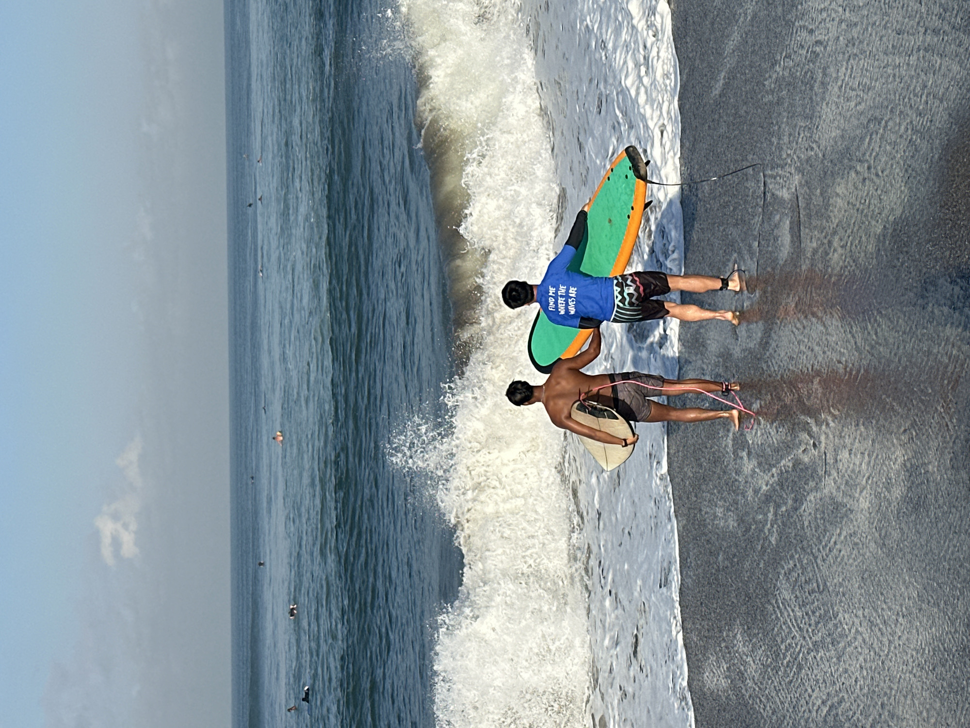 Two individuals, presumed to be surfers, stand at the water's edge on a dark sand beach in North Kuta, Indonesia, holding surfboards. The person on the left has a light-colored surfboard and is wearing dark swim shorts. The person on the right wears a blue t-shirt with white text that reads "FIND ME WHERE THE WAVES ARE" and striped swim shorts, holding a brightly colored green and orange surfboard. A large wave is breaking in the background, creating significant white foam and spray. The ocean extends to the horizon, where it meets a hazy sky with scattered clouds. Other surfers and surfboards are visible in the distance on the water. The lighting suggests it is daytime, likely morning or late afternoon.