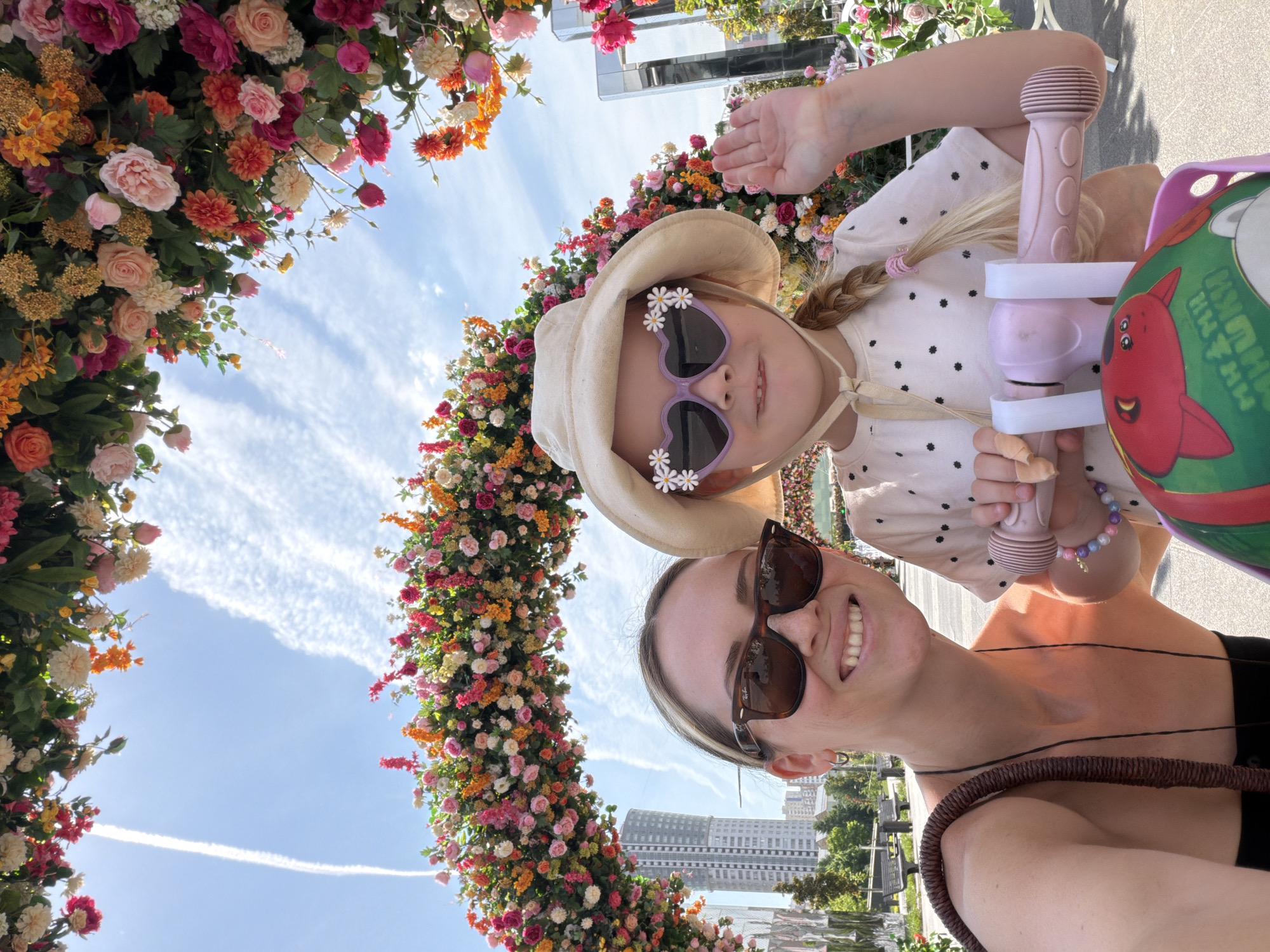 A selfie-style photograph captures an adult female and a young girl standing beneath a large, arching floral display on a bright, sunny day in Khimki, Russia. The floral arch is densely decorated with various colorful blooms, including roses, dahlias, and smaller flowers in shades of pink, orange, red, yellow, and white, interspersed with green foliage. The adult, positioned on the left, smiles directly at the camera, wearing dark tortoiseshell Ray-Ban sunglasses, small silver hoop earrings, and a dark, off-shoulder top with a brown woven bag strap visible over her right shoulder. The young girl, on the right, wears a light-colored bucket hat with a chin strap, distinct heart-shaped purple sunglasses embellished with white daisy cut-outs, and a light-colored t-shirt with dark polka dots. Her blonde hair is styled in a braid secured with a pink elastic band. She raises her left hand in a waving gesture while gripping the pink handlebars of a child's scooter or tricycle. A green basket, decorated with red cartoon characters and partial Cyrillic text, is attached to the front of the scooter. The background features a clear blue sky with some wispy white clouds and a prominent aircraft contrail stretching vertically on the left. In the distance to the left, tall urban buildings, including a prominent high-rise, are visible amidst trees and what appears to be a paved park area with benches. Reflective structures and additional floral arrangements are discernible on the right side of the background.