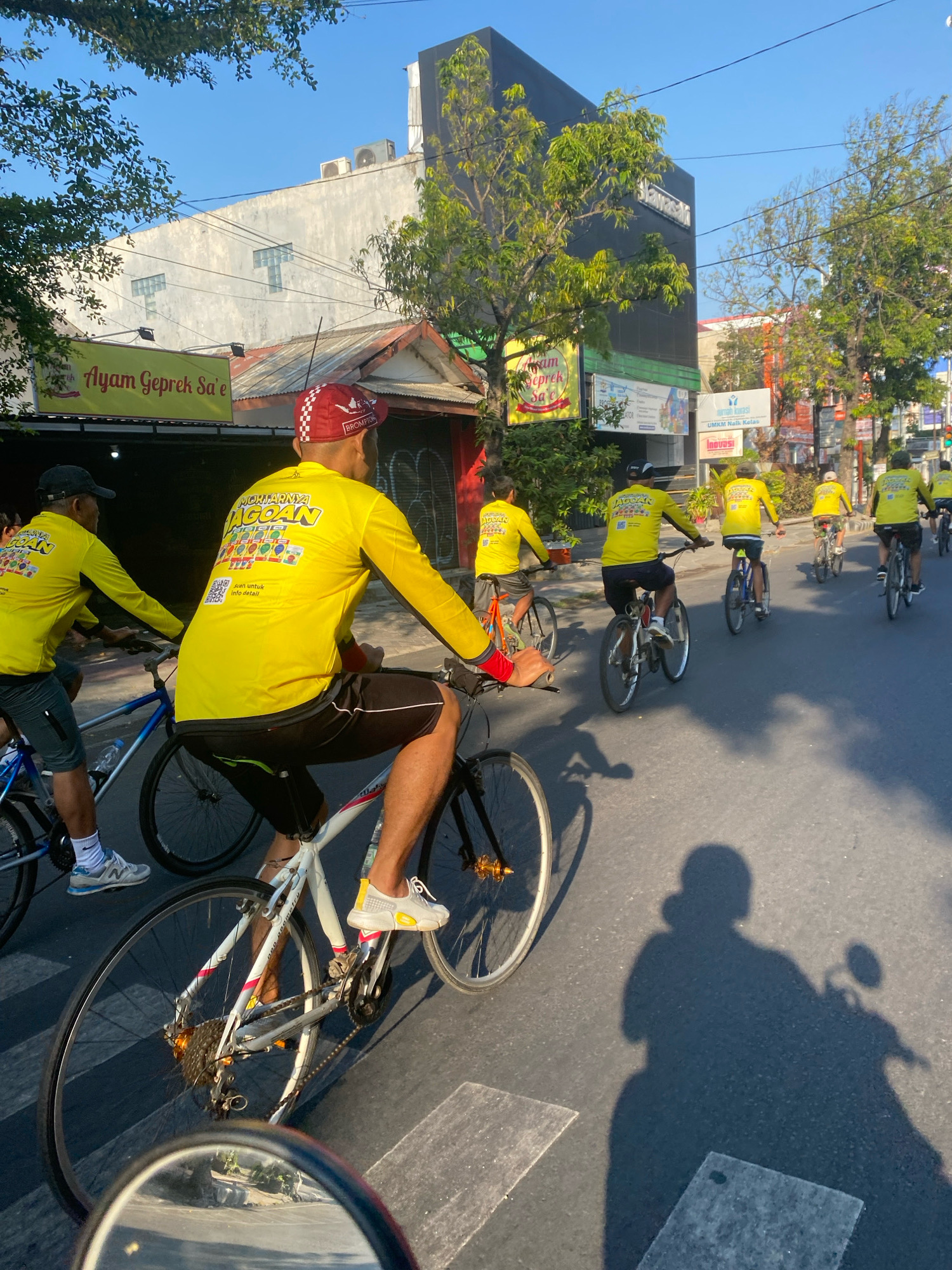 A group of cyclists wearing matching bright yellow long-sleeved shirts and black shorts are riding their bicycles on a paved street, suggesting an organized event or tour in the city of Kediri, Indonesia. The cyclist in the foreground, with his back to the camera, is wearing a red and white checkered cap with a "Brompton" logo. He is riding a white bicycle with red accents. Behind him, other cyclists in similar yellow attire are visible, some on blue bicycles and others on white bicycles. One cyclist is wearing a black cap. The street appears to be a typical urban road with buildings on either side. A sign for "Ayam Geprek Sa'e" is visible on the left, indicating a food establishment. Trees line the street, and the sky is clear and blue, suggesting it is daytime. The cyclists are casting shadows on the asphalt. A partial view of a mirror, likely from a motorcycle, is in the lower left corner of the image, reflecting some of the surroundings.