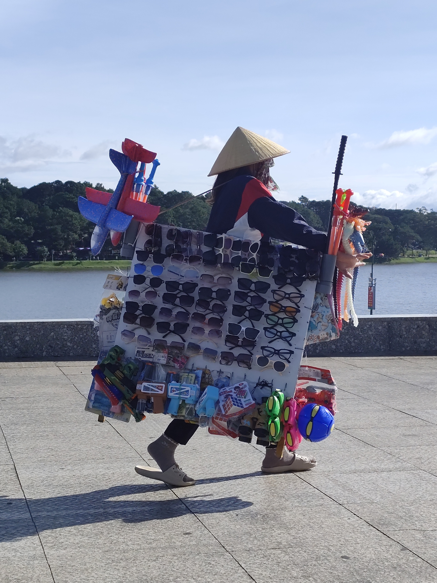 A person wearing a conical hat and a jacket walks along a paved walkway near a body of water, carrying a display of goods. The walkway is made of light gray square tiles. A low, stone wall with a textured surface runs along the edge of the walkway. Beyond the wall is a large body of water, possibly a lake, with a tree-covered hillside in the distance. The person is laden with a mobile display of merchandise, including many pairs of sunglasses, several toy airplanes, toy guns, colorful plastic toys resembling noodles, and a blue ball. The person is wearing dark pants, gray socks, and light-colored slip-on shoes. The conical hat suggests a Southeast Asian context, and the abundance of toys and sunglasses indicates a vendor selling to tourists. The scene appears to be in Da Lat, Vietnam, a city known for its lakes and natural beauty, often frequented by tourists. The sky is bright blue with scattered white clouds. The lighting suggests it is daytime.