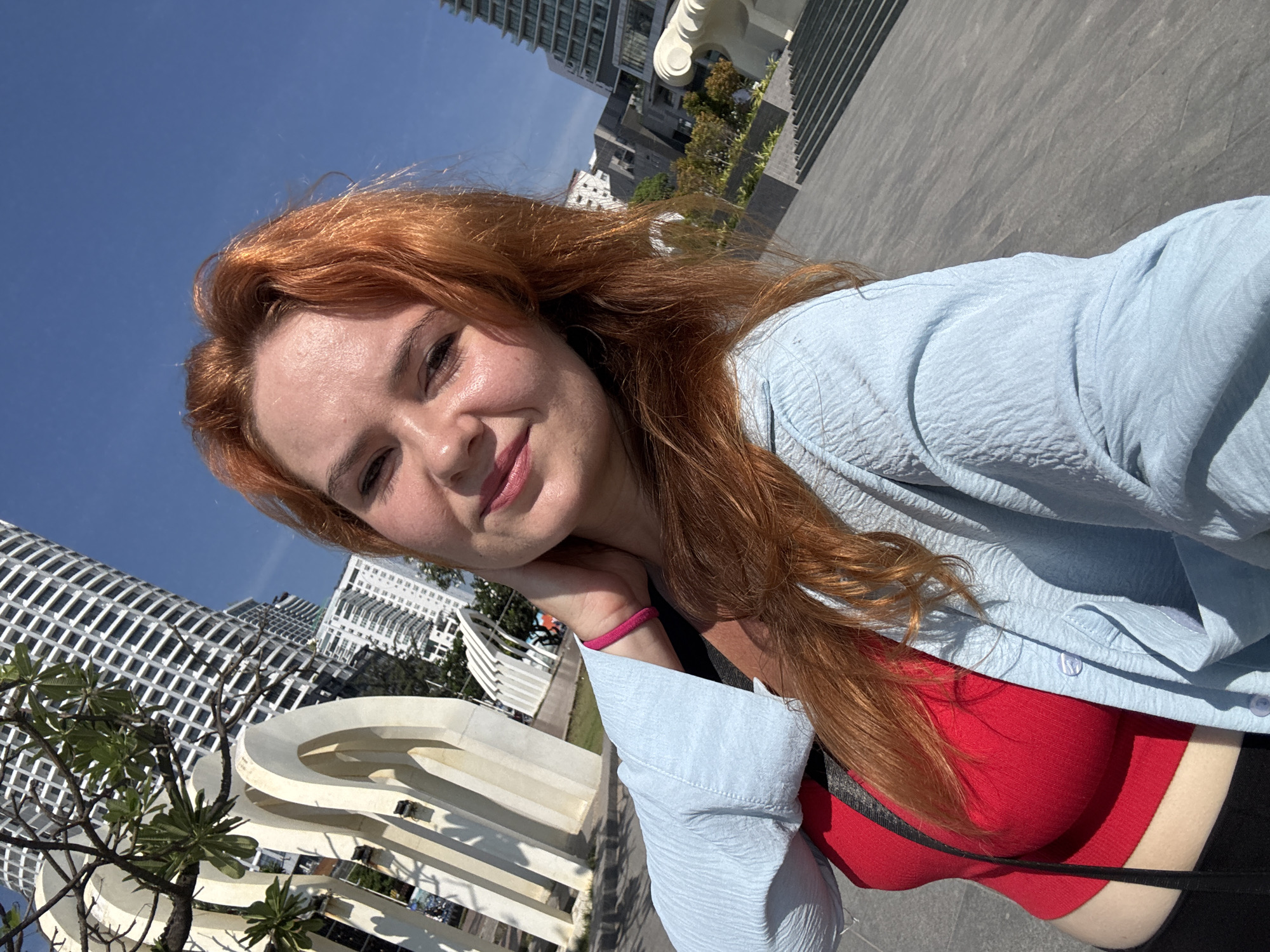 In Nha Trang Ward, Vietnam, a fair-skinned woman with long, bright red hair takes a selfie outdoors on a sunny day. She is wearing a red tank top under a light blue, textured button-down shirt. Her right hand is raised to her neck, and she is smiling at the camera. Behind her, modern high-rise buildings dominate the skyline, with a distinctive white sculptural art piece in the mid-ground and a paved plaza in the foreground. The sky is clear and blue.