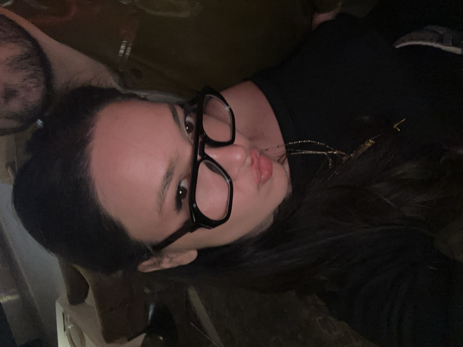 The image features a close-up portrait of two individuals in an indoor, dimly lit setting.
**People:**
1. **Primary Subject (Female):** Positioned centrally and prominently in the foreground, facing the camera. She appears to be an adult with long, dark hair, parted on the side. She wears dark-rimmed glasses, obscuring part of her eyes. Her lips are pursed in an expressive gesture. She is wearing a dark, likely long-sleeved, top or shirt. Multiple thin, gold-colored necklaces are visible draped over her chest.
2. **Secondary Subject (Male):** Partially visible on the right side of the frame, behind and to the right of the primary subject. Only his upper chest, right shoulder, and the lower portion of his face (including a dark beard) are visible. He appears to be wearing a dark, possibly olive green, jacket or shirt.
**Objects:**
* One pair of dark-rimmed glasses with a double bridge, worn by the female subject.
* Several thin, gold-colored necklaces with small charms or pendants, worn by the female subject.
**Actions:**
* The female subject is looking directly at the camera with a distinct facial expression involving pursed lips.
* The male subject's visible portion suggests he is positioned closely behind the female subject.
**Environment:**
* The background is dark and out of focus, indicating an indoor location with low ambient light.
* Indistinct shapes, possibly furniture or decorative elements, are faintly visible in the upper left background, suggesting a room interior.
* The lighting is soft and appears to be artificial, likely originating from above or to the front of the subjects.
**Location:**
The image contains no identifiable visual markers (e.g., landmarks, street signs, architectural styles specific to Yerevan) to confirm the city of Yerevan, Armenia. Therefore, the precise location within Yerevan cannot be determined from the image data provided.