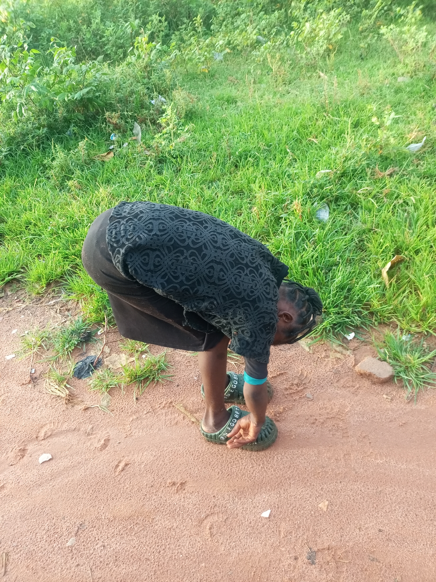 A young Black person is bending over, facing down, and reaching towards the ground. They are wearing a black patterned top and a black skirt. On their feet are green slide sandals with "EGO BETTER" visible on the straps. A teal blue band is worn around their left wrist. The person is positioned on a reddish-brown dirt path with sparse green grass growing at its edges. In the background, there is a dense area of lush green vegetation, suggesting an outdoor, possibly rural, setting. Some scattered white and grey debris is visible in the vegetation. The lighting indicates it is daytime, and the environment appears dry. The overall scene suggests the person may be picking something up from the ground or examining something. The location is specified as Tempure, Nigeria.