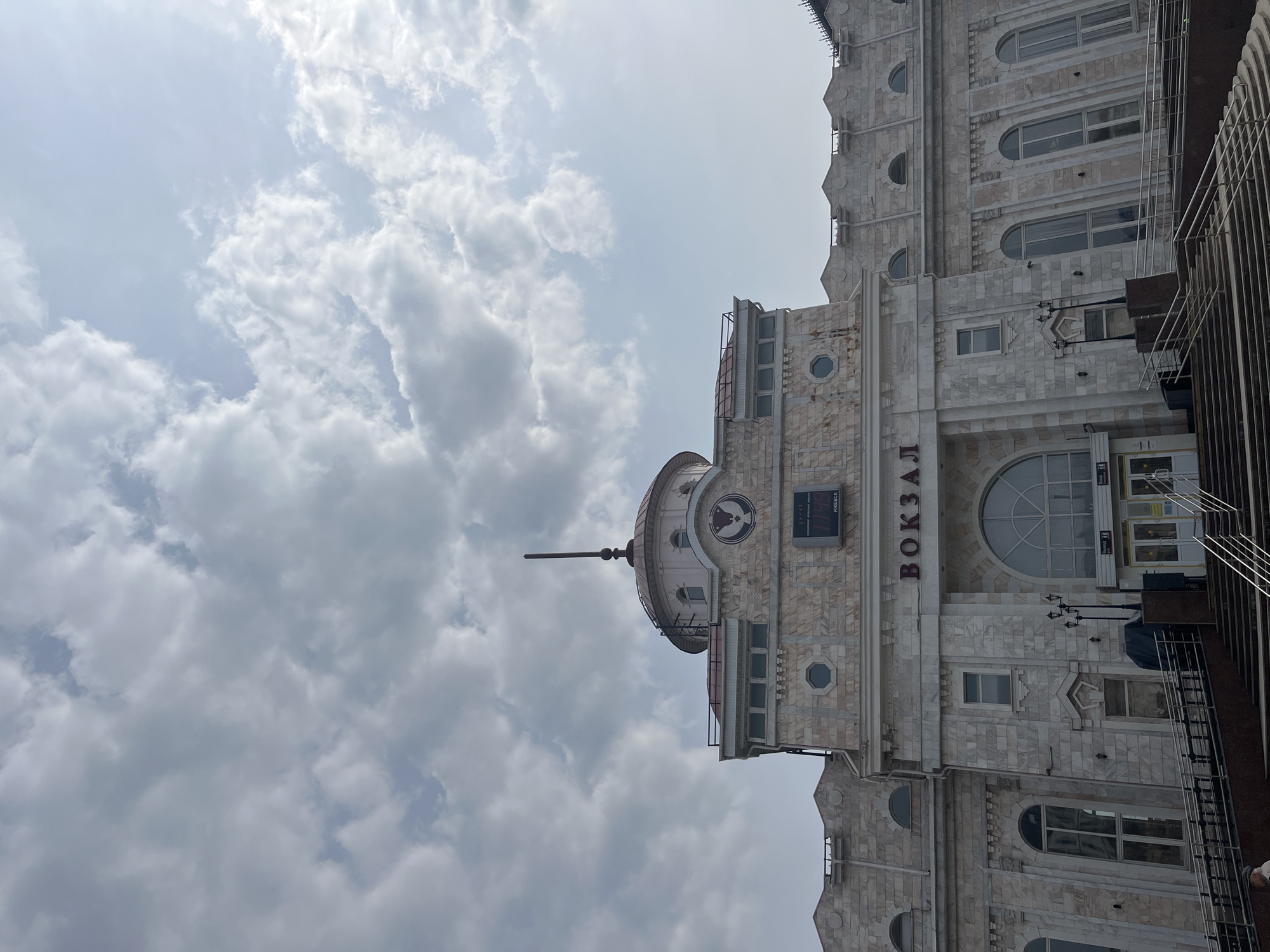 The image shows the Izhevsk railway station in Russia, viewed from a low angle, making it appear tall. The building is made of light-colored stone, with architectural details such as arched windows, small circular windows, and a dome-shaped tower with a spire. The name of the station, "ВОКЗАЛ" (Vokzal), is displayed in Cyrillic letters. The sky is filled with heavy clouds, suggesting overcast weather. Part of a metal structure is visible on the right side, slightly obscuring the view of the building.