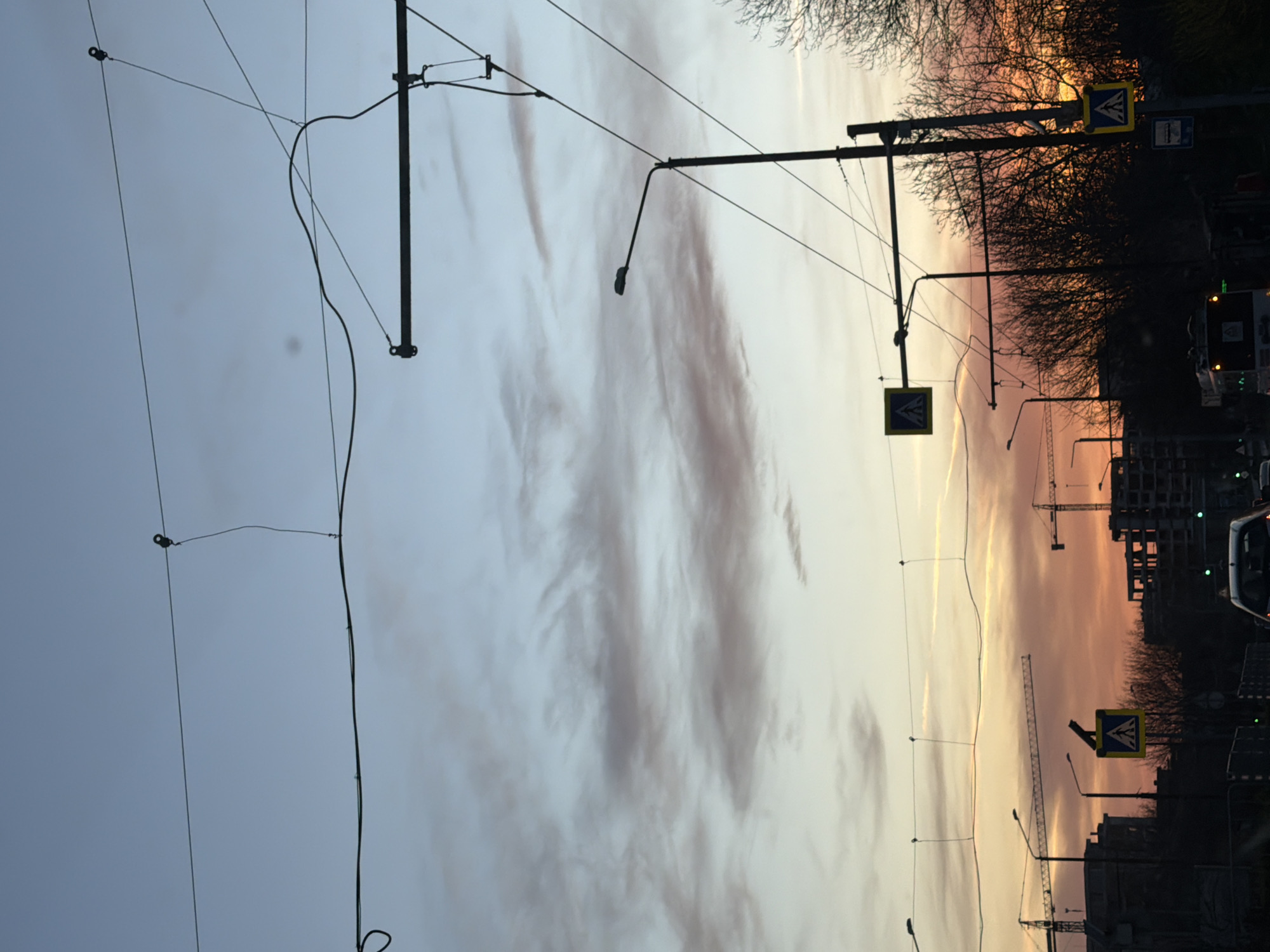 The image depicts an urban scene at sunset in Varna, Bulgaria, characterized by a mixture of infrastructure and construction. The sky transitions from a pale blue to a warm orange with streaky clouds, suggesting a clear evening. Overhead, there are tram wires and supporting structures extending across the scene, typical of urban transportation systems. Streetlights are positioned along the road, silhouetted against the fading sunlight. The foreground reveals pedestrian crossing signs and infrastructure. In the distance, the silhouettes of buildings under construction are visible, with at least two cranes extending into the sky, indicating ongoing development. The presence of trees lining the street provides an element of greenery. The photograph appears to have been taken from a slightly low angle, possibly from within a vehicle or at street level.