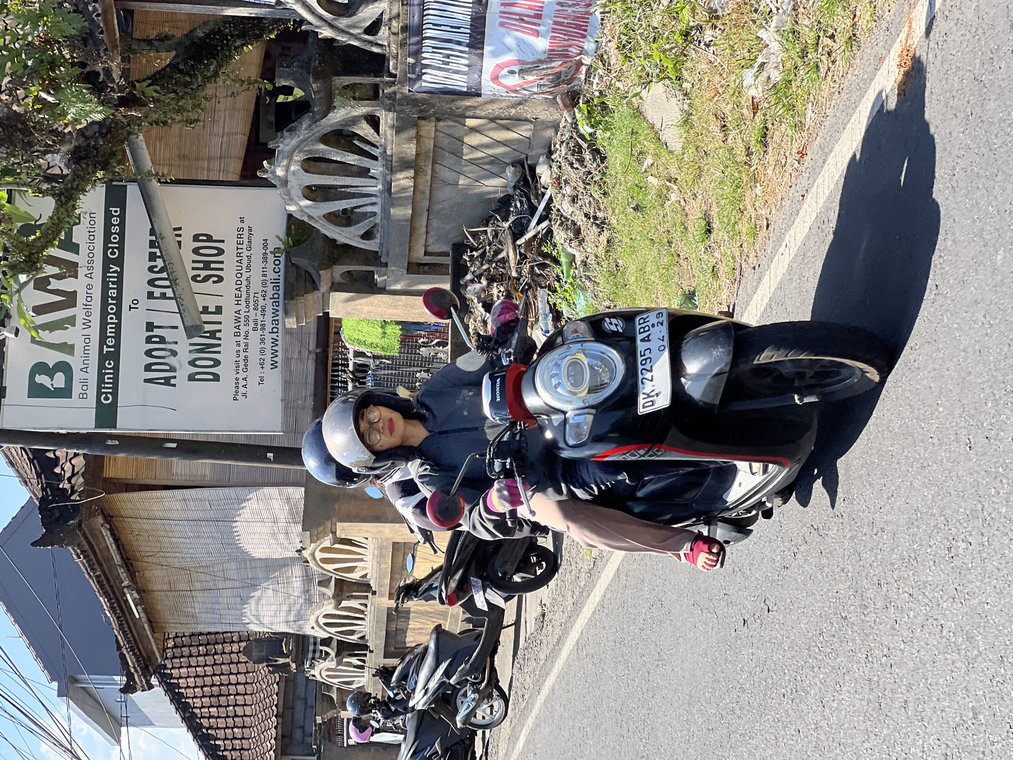 In the city of Ubud District, Indonesia, a person wearing glasses and a grey hoodie rides as a passenger on a black scooter. The driver of the scooter is partially visible. Both individuals are wearing helmets. The scooter has a license plate with the inscription "DK 2295 ABR 04.29". The scooter is traveling on a paved road. In the background, there are buildings with traditional Balinese architecture, including tiled roofs and woven bamboo shades. A sign visible in the background reads "BAWA Bali Animal Welfare Association", indicating a local animal welfare organization. The surrounding environment appears somewhat cluttered, with a pile of trash and debris visible near a wall. The scene is illuminated by bright daylight.