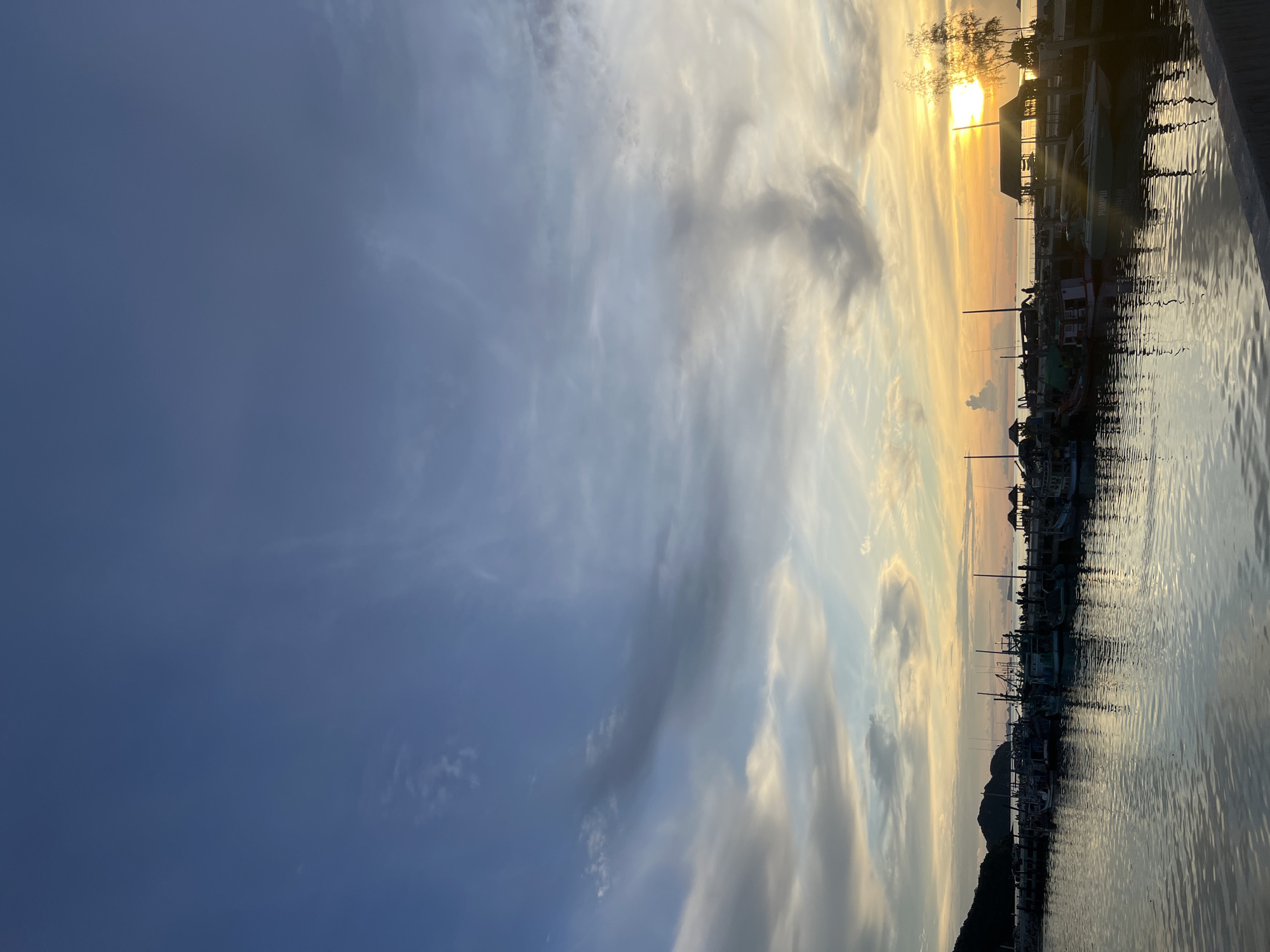 The image depicts a serene harbor scene at sunset, likely in a coastal city. The sky is a canvas of blues and oranges, with dramatic cloud formations swirling across the horizon. The sun, a brilliant orb, is low in the sky, casting a warm glow and reflecting on the calm waters of the harbor. A line of fishing boats is docked along a pier, their silhouettes stark against the vibrant sky. The boats vary in size and design, some appearing weathered and well-used, others more modern. Small, rustic structures, possibly shelters or small buildings, are visible on the pier, interspersed with the boats. In the background, a distant landmass or island is visible, adding depth to the landscape. The overall atmosphere is tranquil and picturesque, conveying a sense of peace and the end of a day's labor.