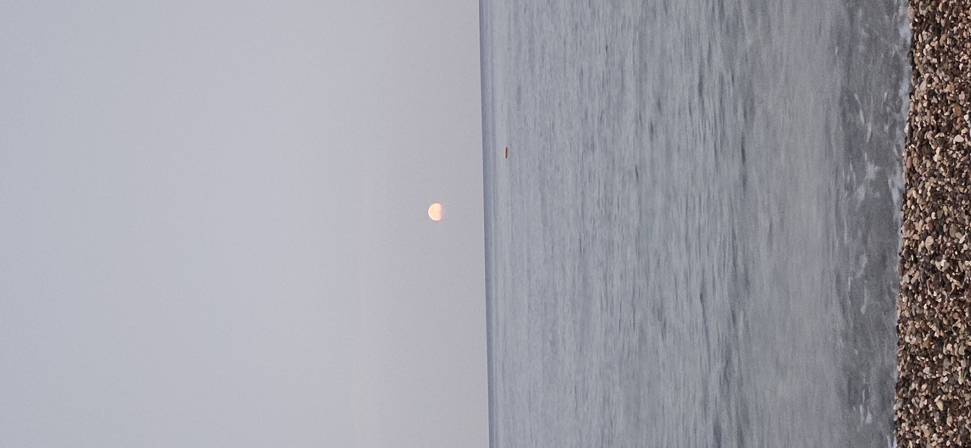 The image captures a serene seascape at dusk, likely in Kemer, Turkey. The horizon is defined by the gentle meeting of the vast, calm sea and a pale, overcast sky. A soft, pale-pink moon, partially obscured and appearing low in the sky, is visible slightly left of the center. In the distance on the water, a small, rectangular yellow object, possibly a buoy or a small flotation device, is present. The foreground is a pebbled beach, with the stones exhibiting a variety of earthy tones, from light beige to dark brown and grey. The water in the foreground is shallow and clear, revealing the pebbles beneath the surface, with gentle ripples breaking on the shore. The overall impression is one of quiet tranquility, characteristic of a peaceful evening by the Mediterranean coast.