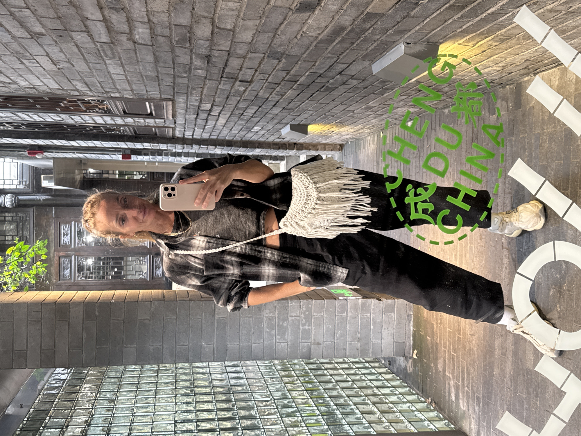 A person is holding a cell phone in their left hand, appearing to take a selfie, in a street-level hallway in the Qingyang District, Chengdu, China. The person is wearing a grey and black plaid shirt over a dark grey top, black pants, and white sneakers. They have a white fringe purse that hangs over their body. The environment is a narrow brick corridor with a glass block wall to the left of the person. A sign on the wall behind the person, that appears to be on a reflective surface, reads "CHENG DU CHINA" in green lettering. The corridor appears to be well-lit with overhead light fixtures.