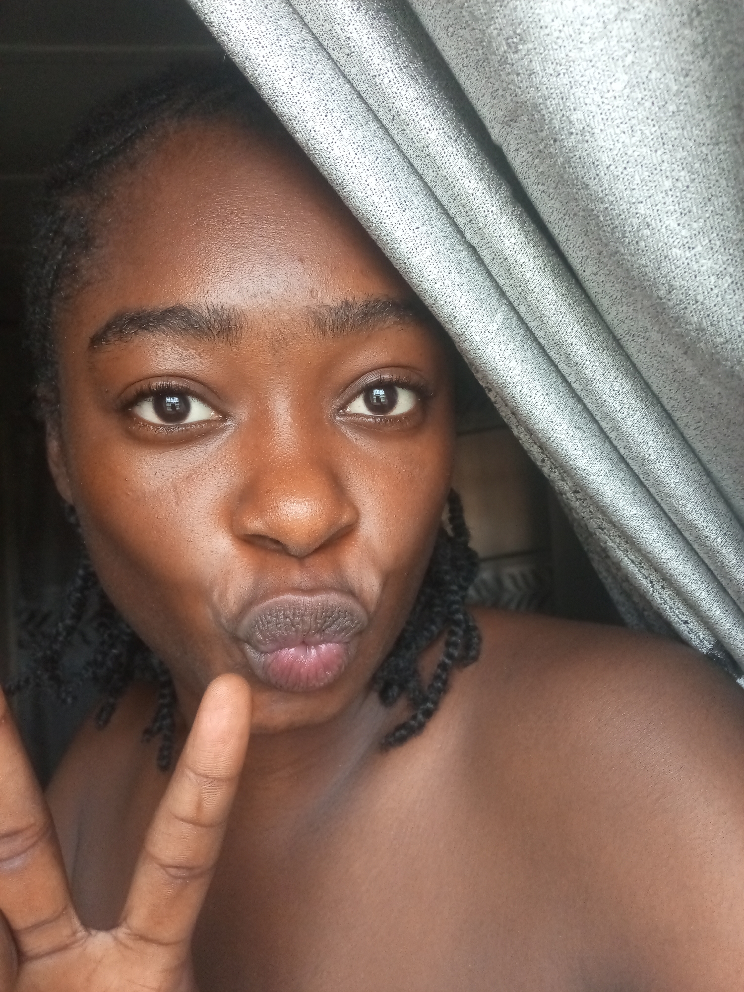 A close-up, waist-up shot shows a young Black woman with dark skin and braided hair. She is facing the camera, with her lips pursed and her left hand in a peace sign gesture near her face. A portion of a grey, textured curtain is visible on the right side of the frame, partially obscuring her shoulder and head. The background is dark and indistinct, suggesting an indoor setting. The lighting is somewhat dim, highlighting the sheen on her skin. The image appears to have been taken in an urban environment, potentially within a home or room in Rafin Par, Nigeria.