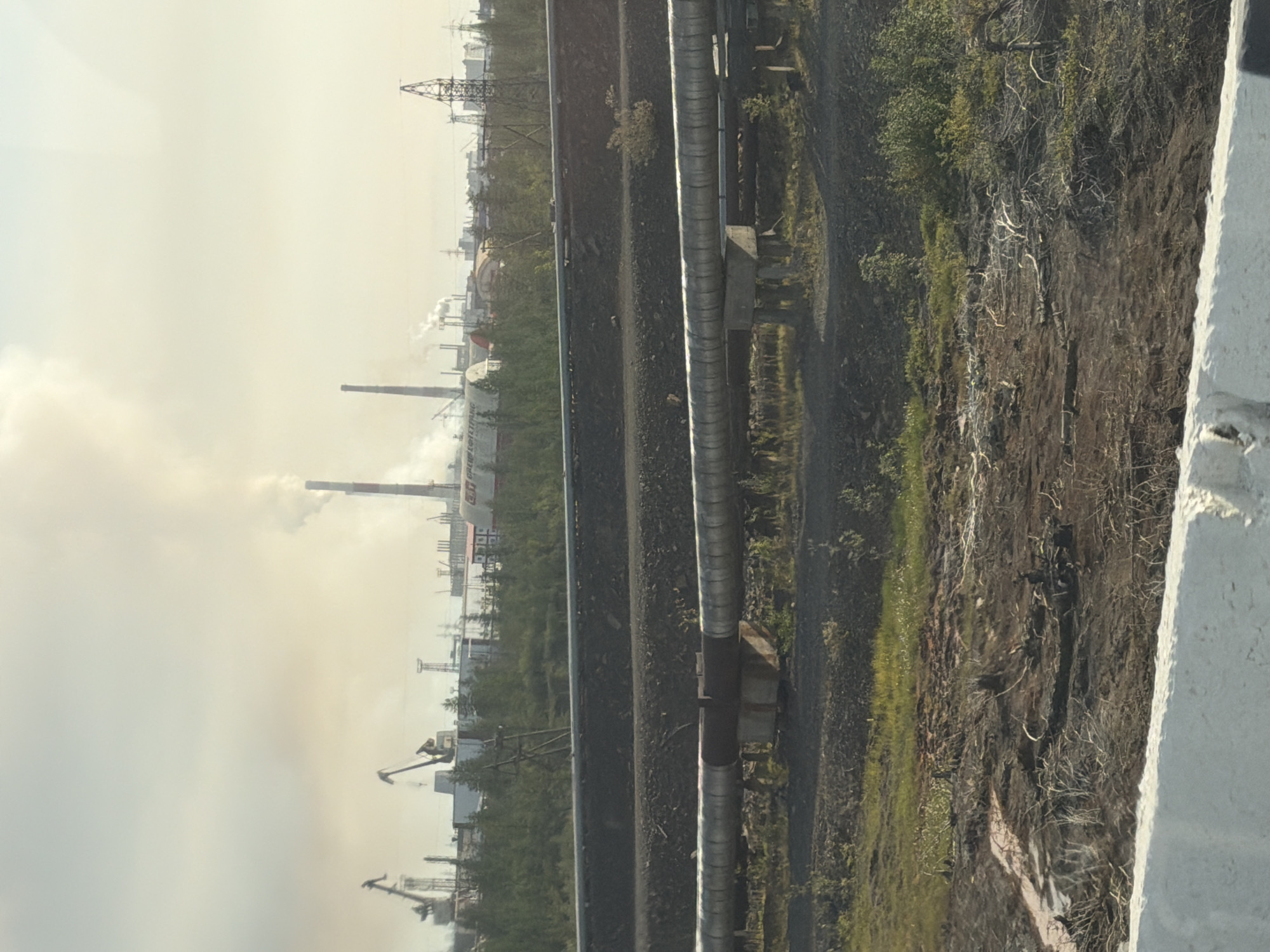 The image depicts an industrial landscape in Norilsk, Russia, characterized by a large factory complex with smoking chimneys, cranes, and industrial structures. In the foreground, several large-diameter pipes, supported by concrete pillars, run horizontally across the frame, indicating an extensive pipeline system. The ground in the foreground is uneven, with patches of dry vegetation, exposed soil, and debris, suggesting a rugged, possibly polluted environment. The background shows a dense line of trees separating the industrial area from the sky, which is hazy with smoke and light from the sun. The overall scene conveys a sense of heavy industrial activity within a vast, open, and somewhat desolate landscape. There are no people visible in the image.